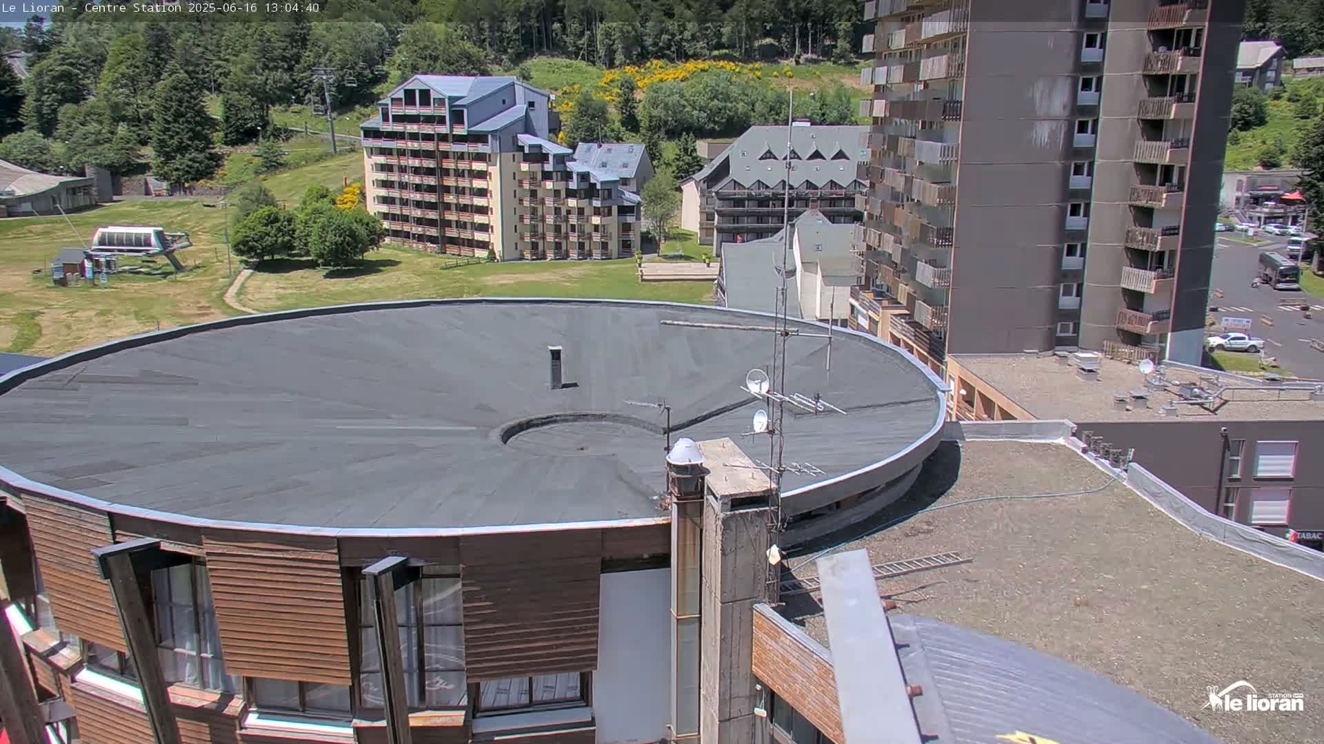 A round building with a dark flat roof is surrounded by other buildings and a grassy area on a sunny day.