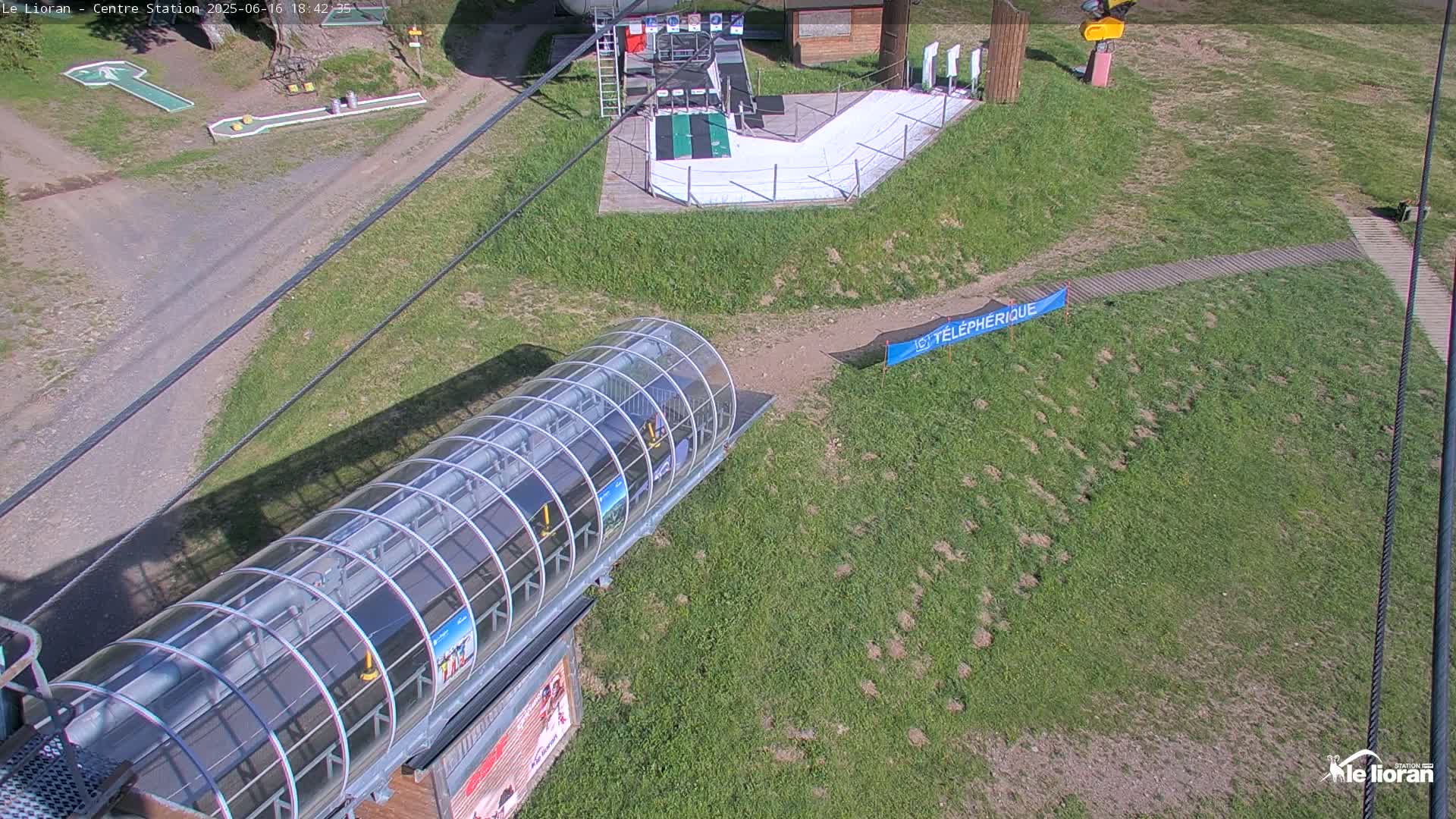 An aerial view shows a grassy area with a covered ski lift, a small snow ramp, and a miniature golf course, under sunny conditions.