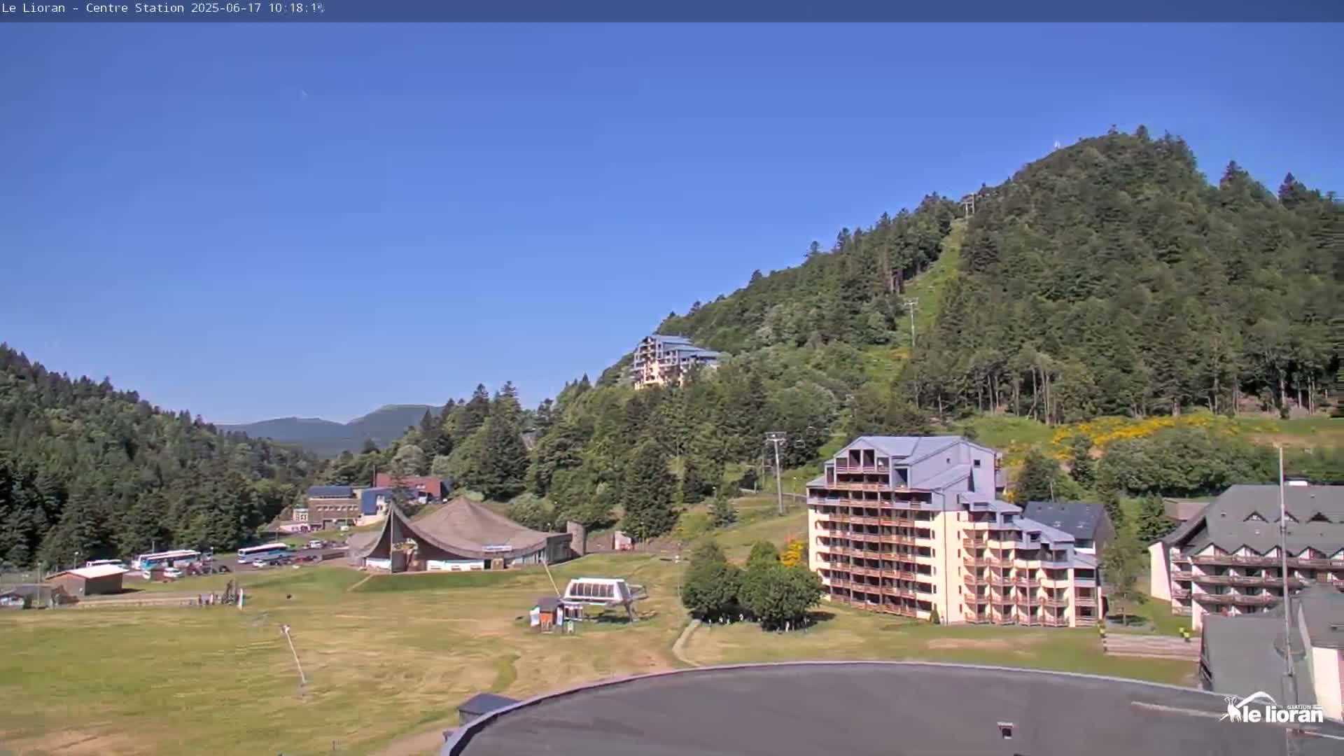 A sunny day in a mountainous region shows several buildings nestled among green hills and trees, with a grassy area in the foreground.