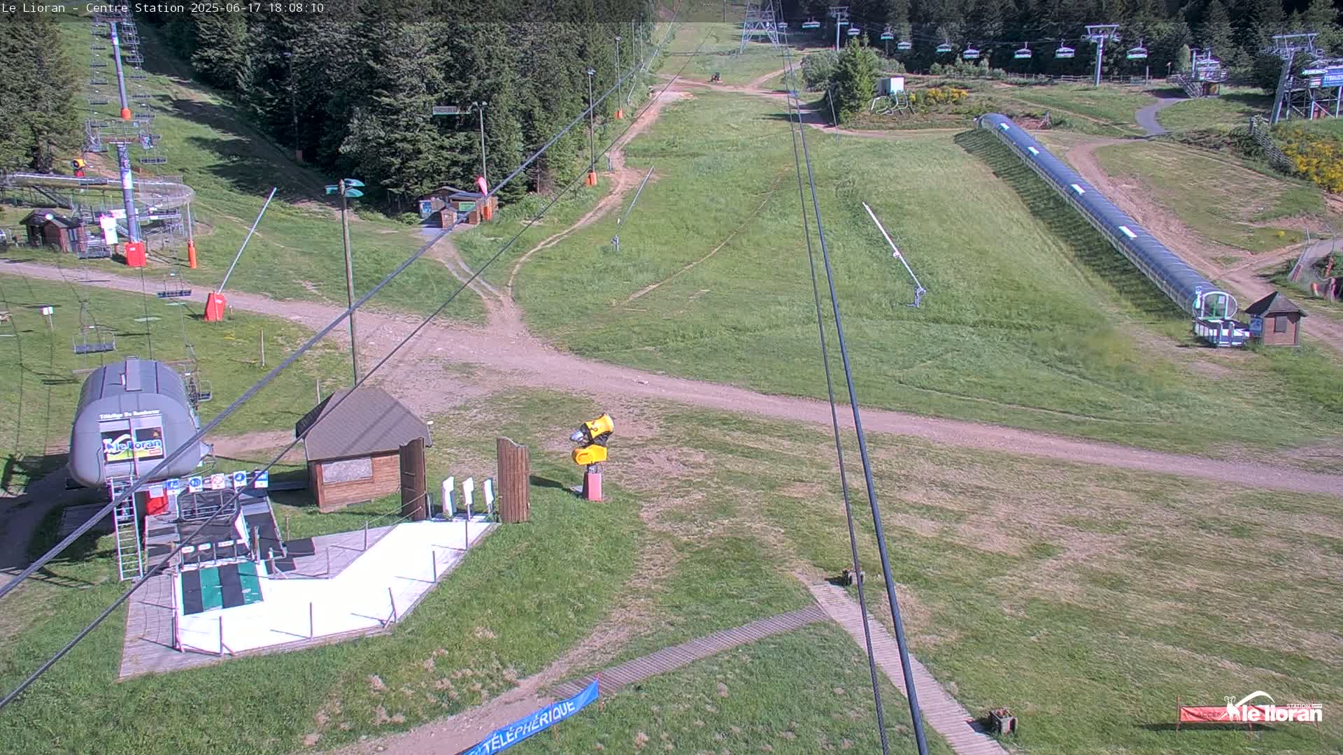 An aerial view of a ski resort on a sunny day shows grassy hills, ski lifts, and a snow-making machine.