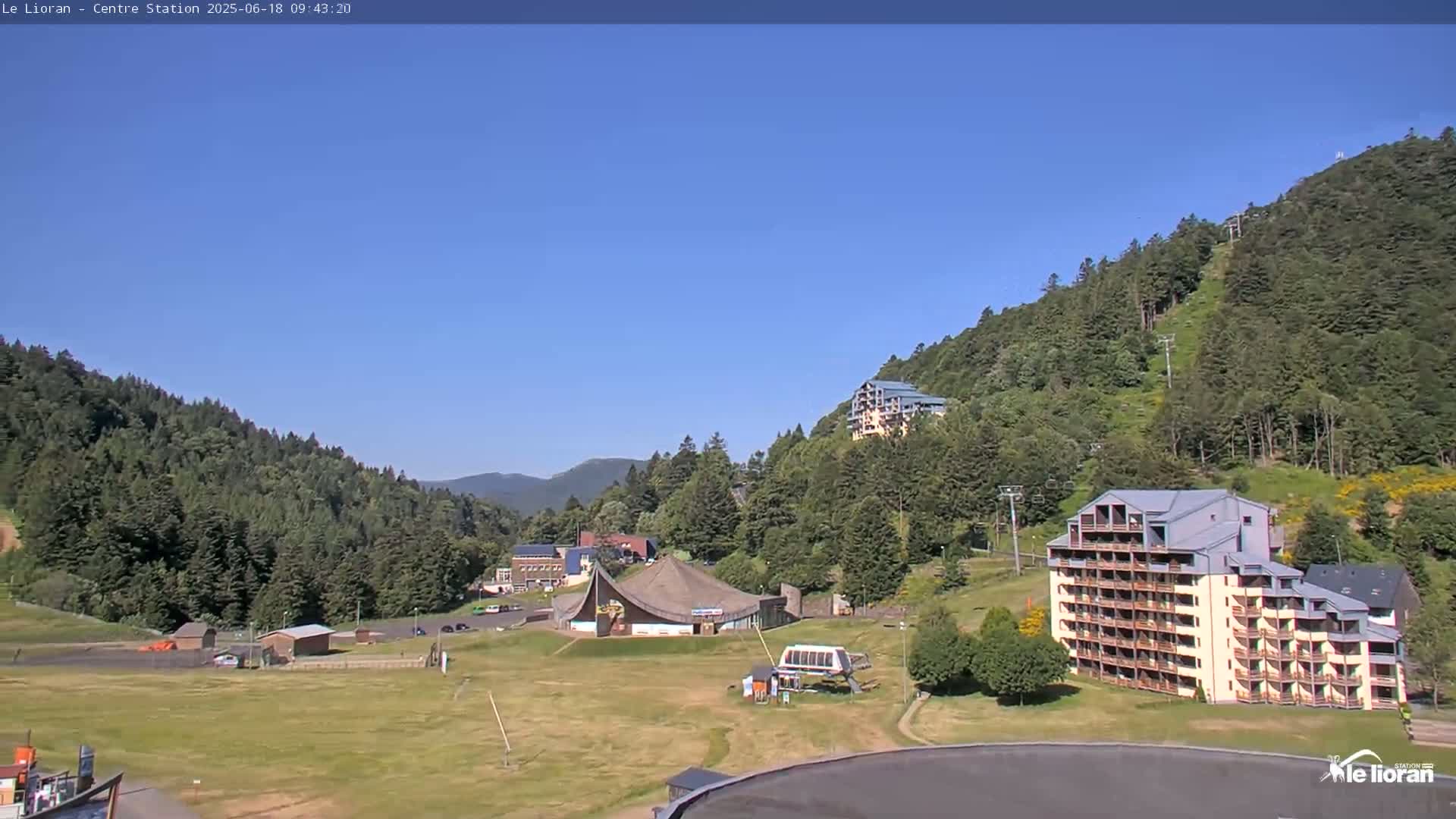 A sunny day reveals a mountainside ski resort with several buildings, a grassy area, and a chairlift, nestled among lush green forests.