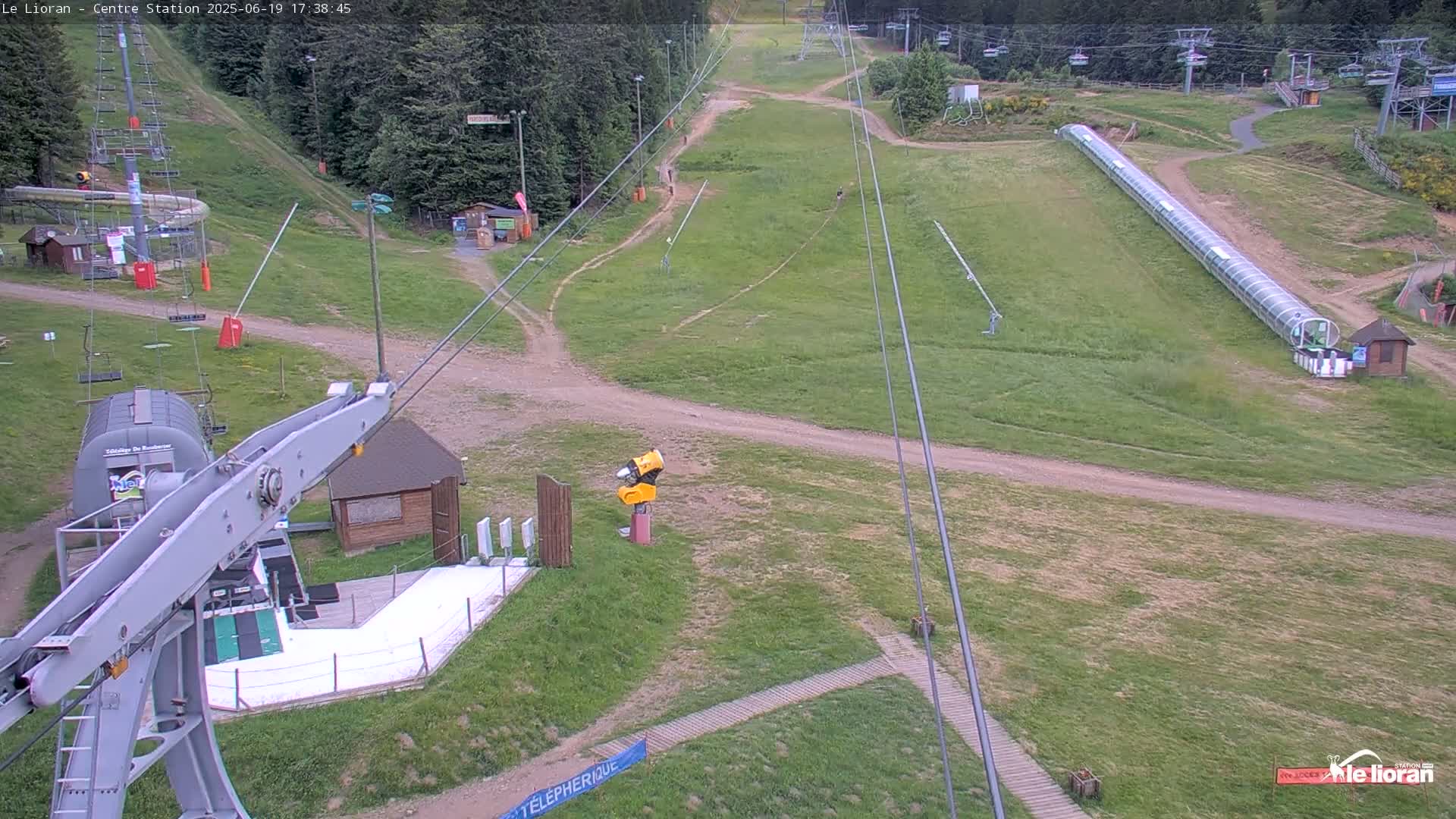 A high-angle view of a ski resort in the daytime, showing grassy hills, ski lifts, and a long, enclosed summer toboggan run.