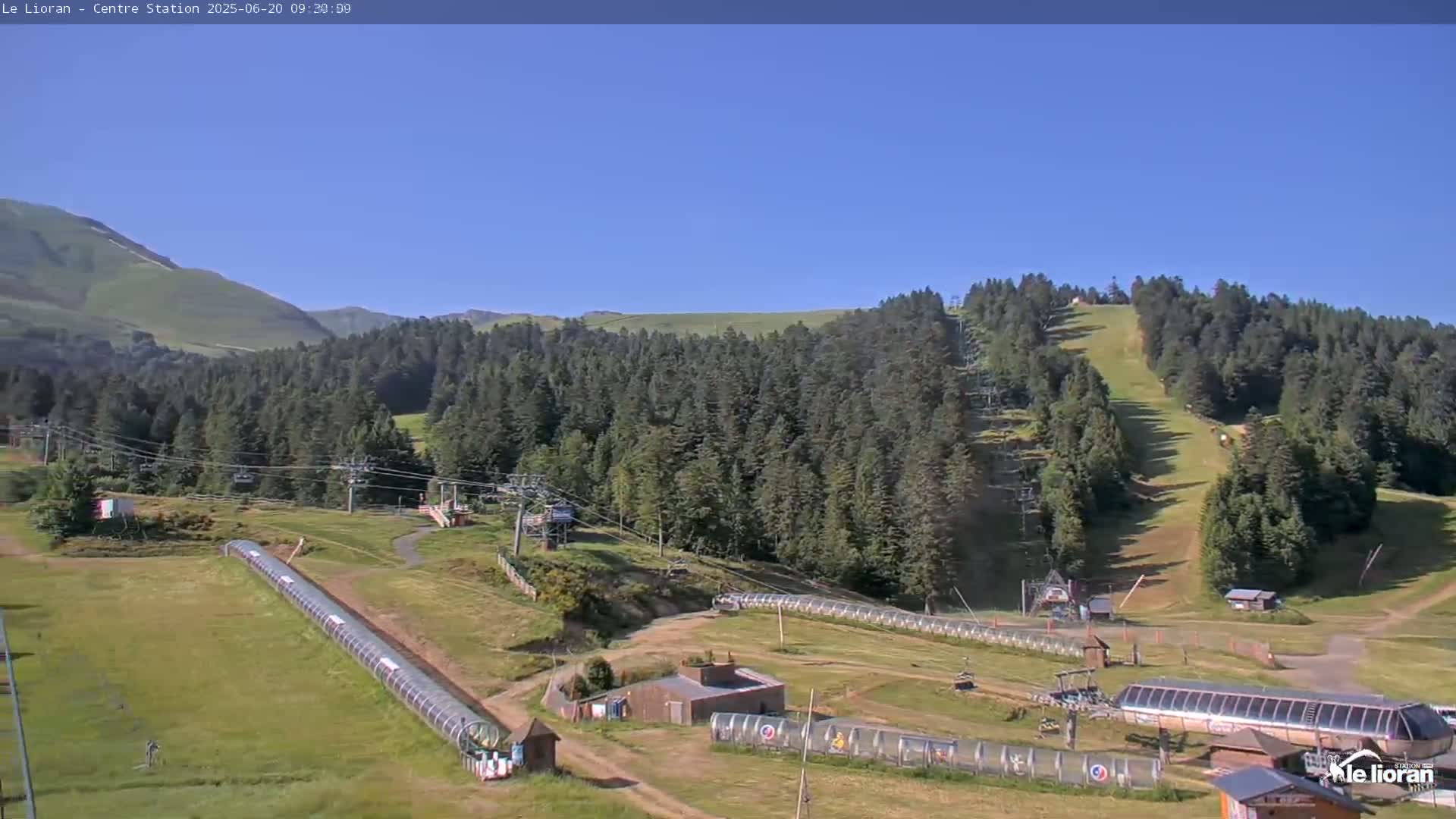 A sunny, clear day reveals a mountainside ski resort with chairlifts, grassy slopes, and long covered conveyors.