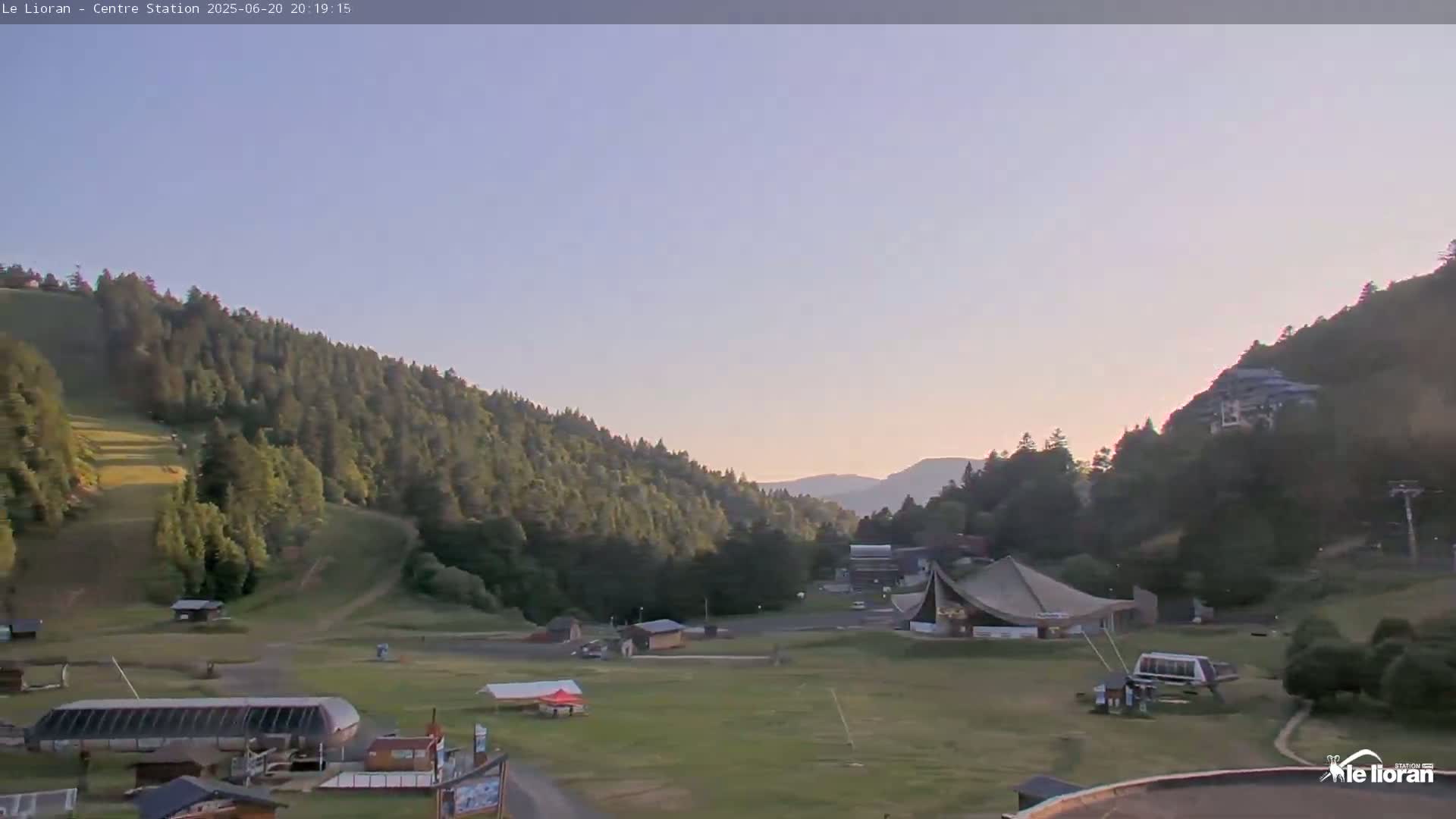 A valley surrounded by green hills and a clear sky at sunset shows a grassy area with several buildings and ski lifts.