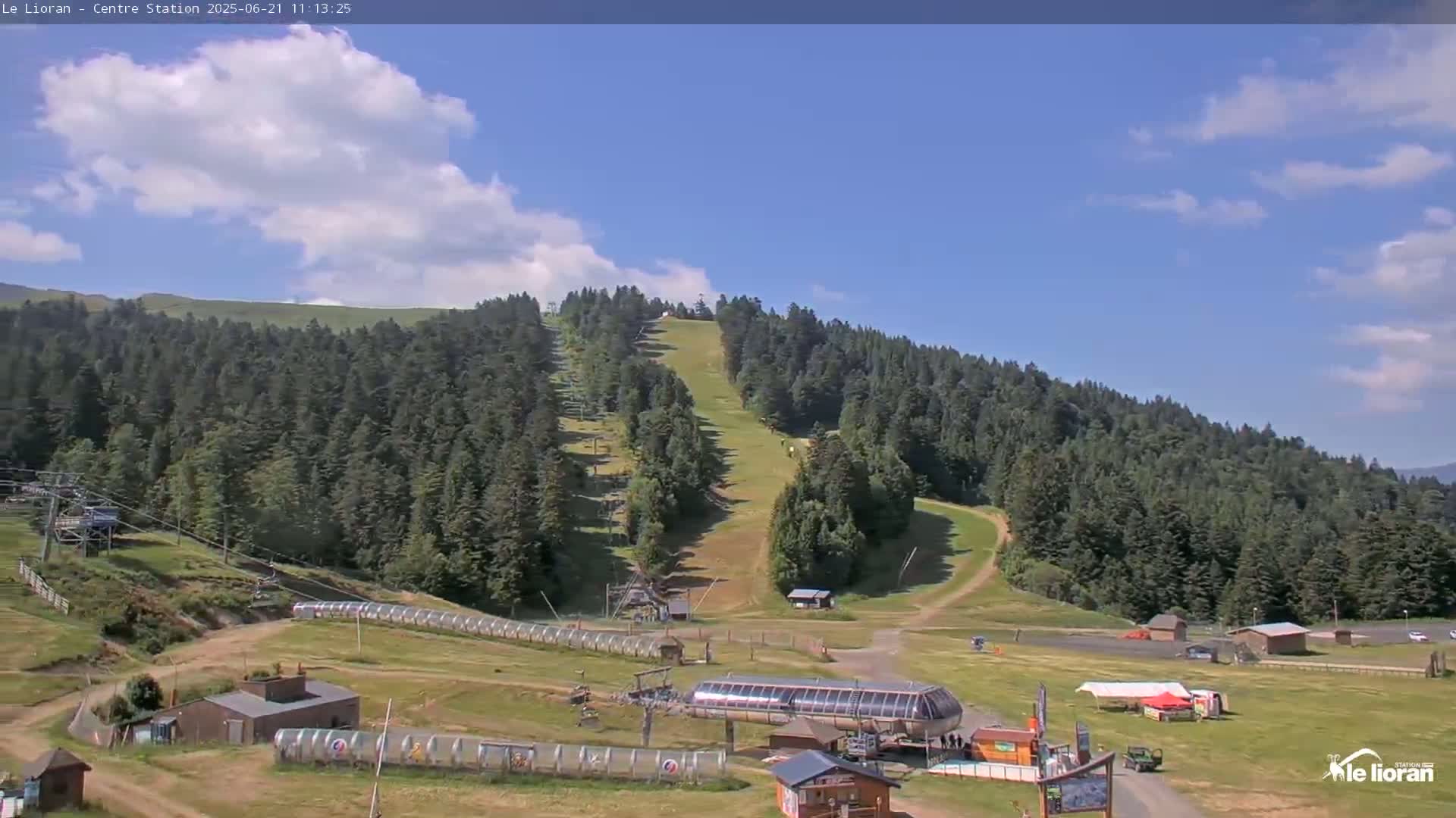 A mostly sunny day reveals a ski resort's mountainside with grassy slopes, ski lifts, and several buildings at the base.