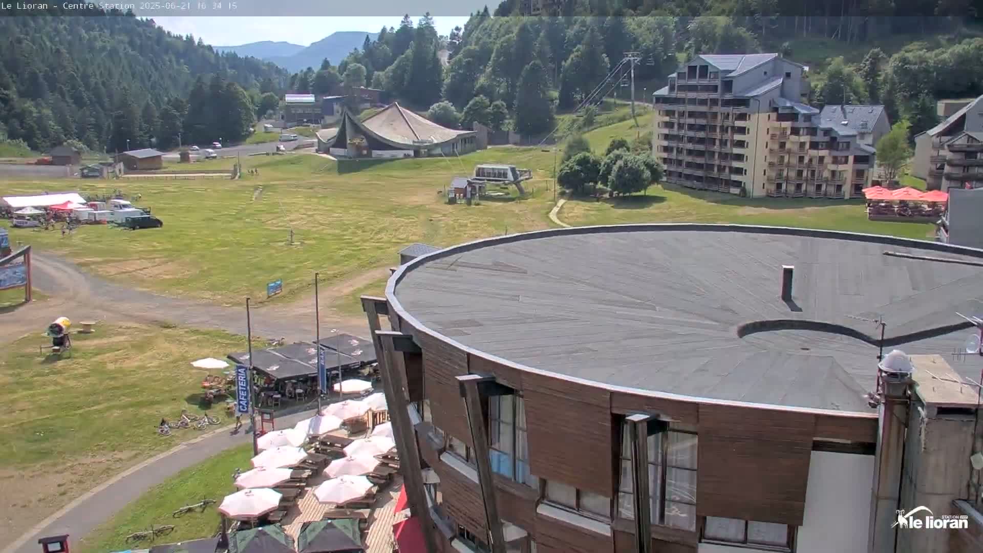 A sunny view of a mountain resort with buildings, a grassy area, and outdoor seating under umbrellas.