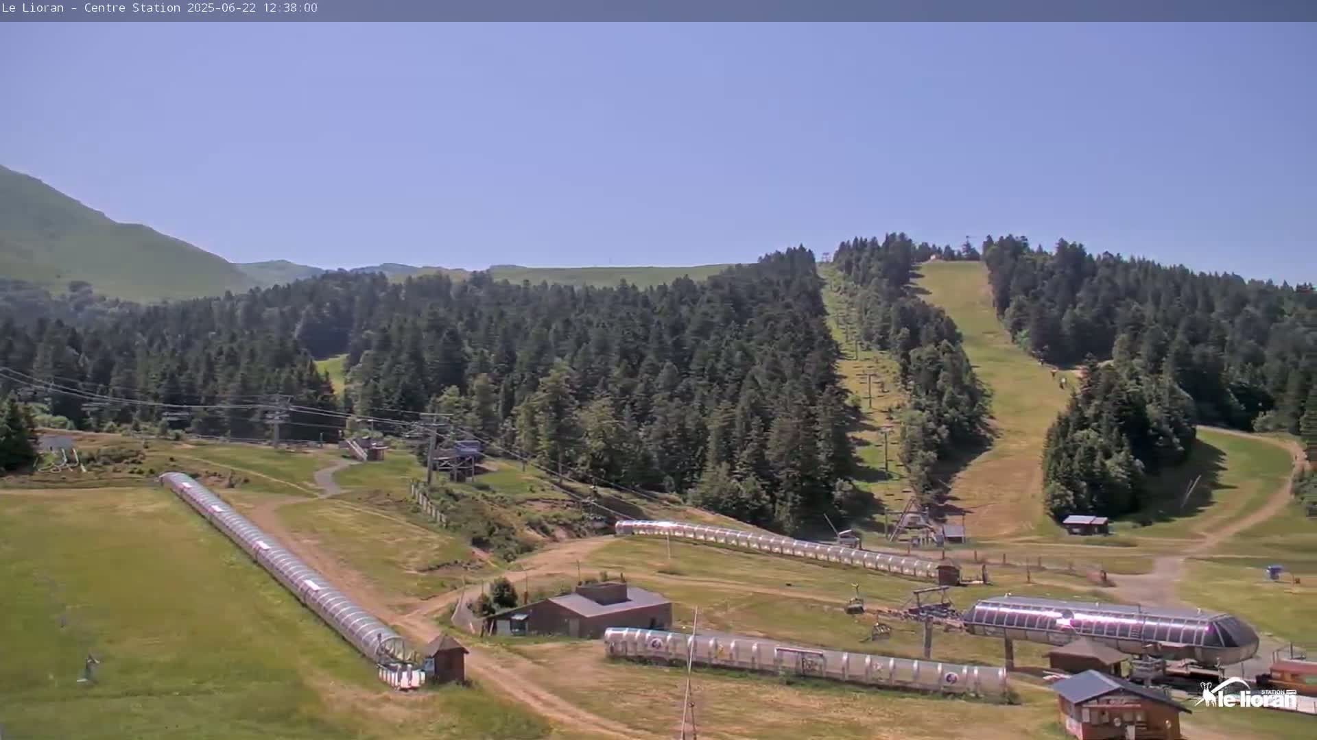 A sunny day overlooking a mountain ski resort with grassy slopes, ski lifts, and covered conveyor belts.