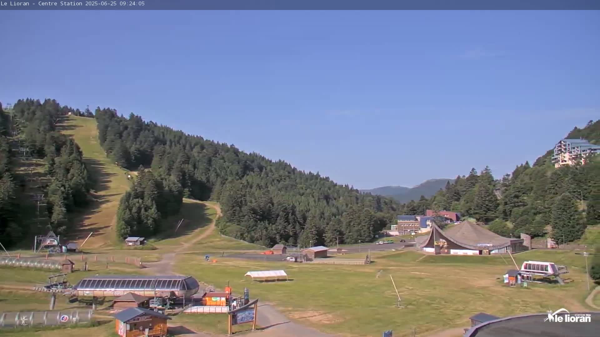 A grassy ski resort area, under a clear blue sky, features a ski lift, several buildings, and a hillside covered in evergreen trees.
