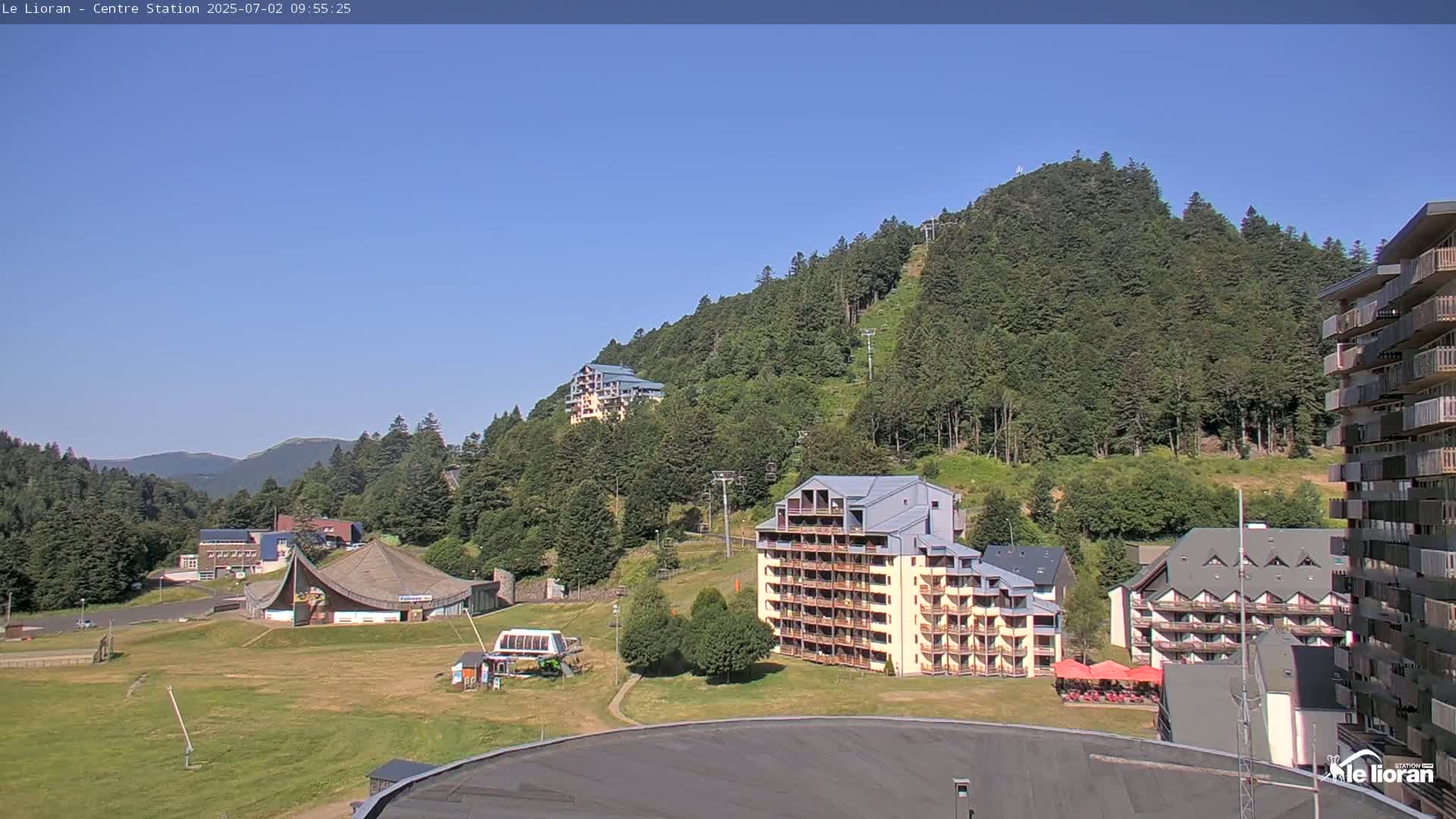 A sunny view of a mountainside resort area with several multi-story buildings, a large grassy area, and ski lift infrastructure.