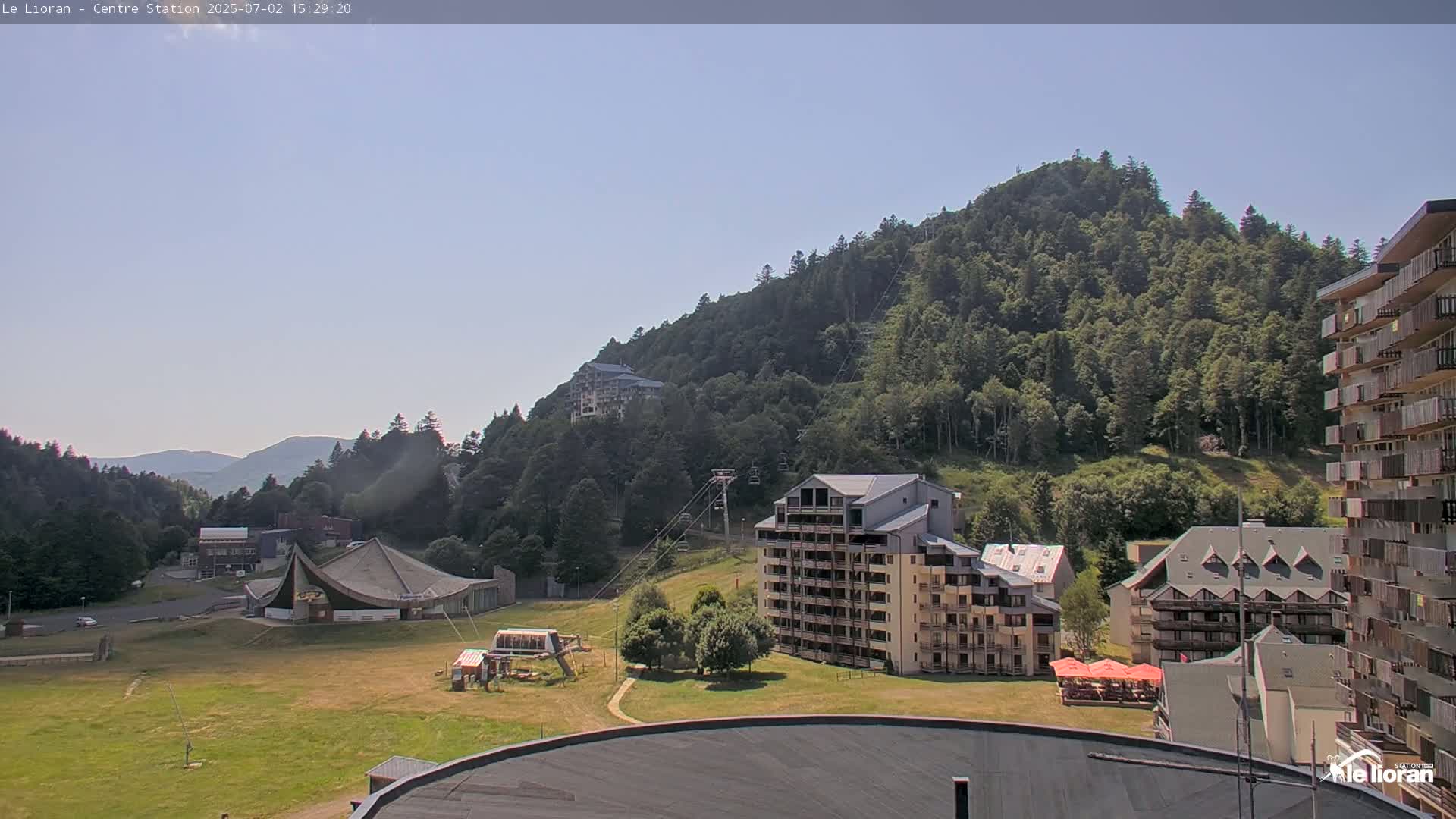 A sunny view of a mountainside resort with several buildings, grassy areas, and a chairlift visible against a clear sky.