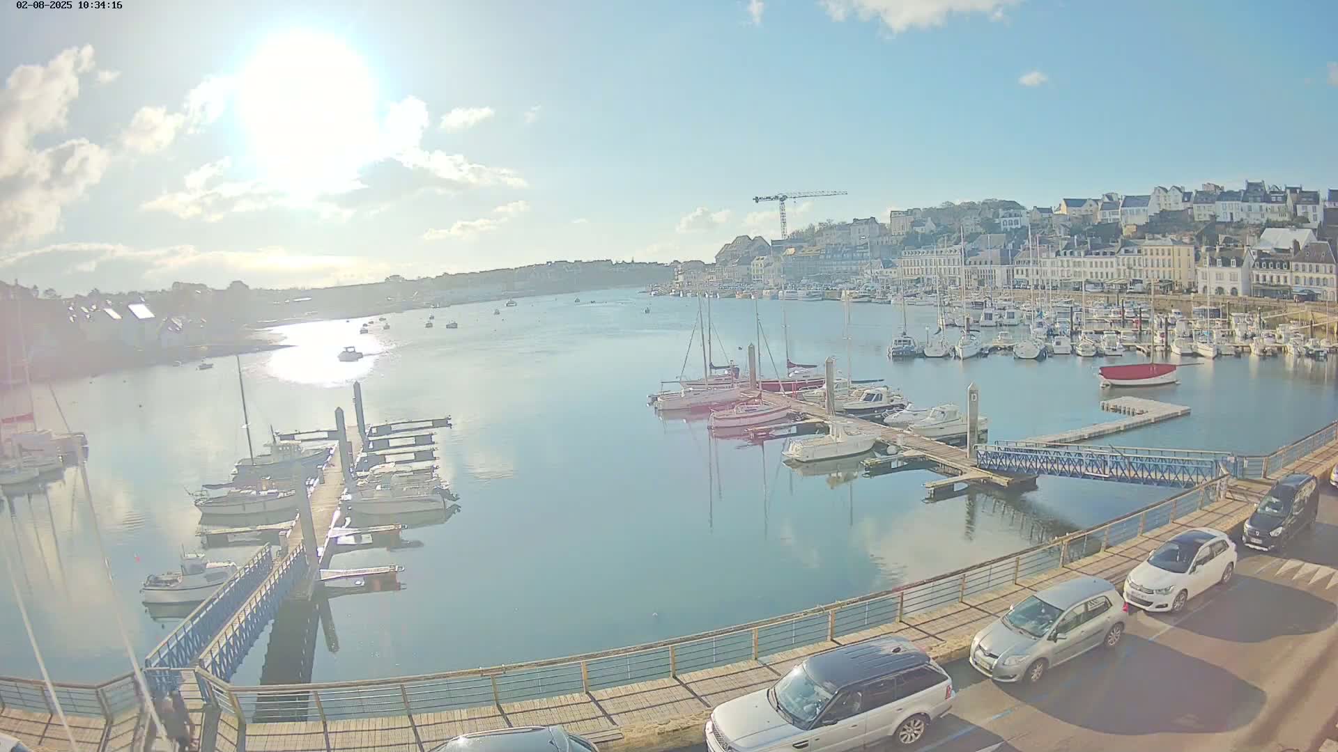 A sunny day shows a harbor filled with boats and yachts docked at piers, with a town in the background and cars parked along a walkway.