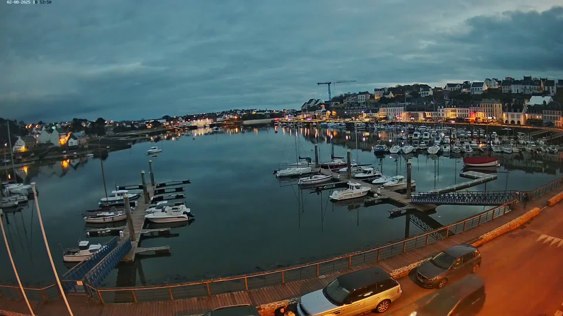 A harbor filled with boats at dusk under a cloudy sky, with cars parked along a nearby roadway.