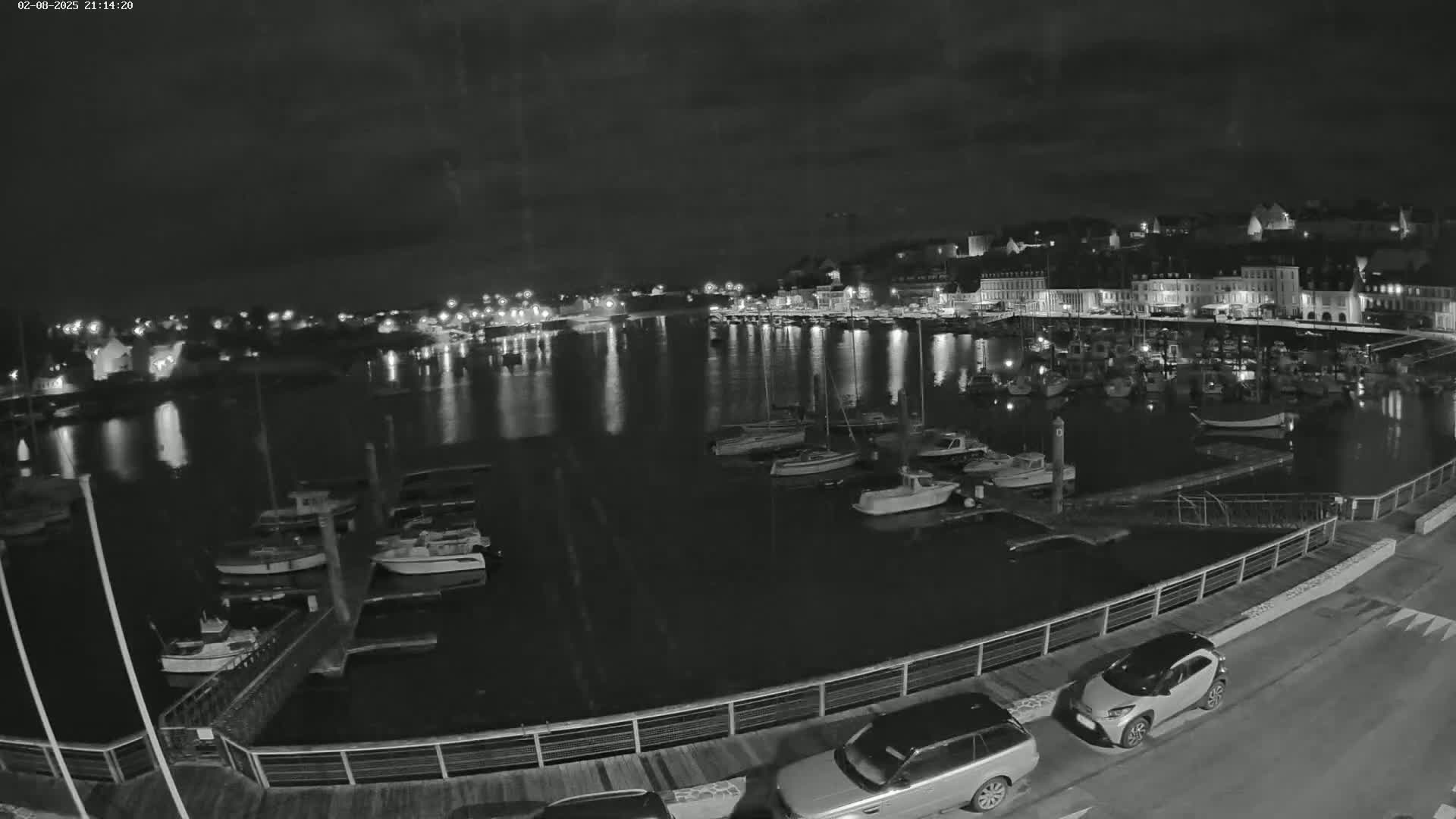 A nighttime view of a harbor filled with boats, a town with lights visible across the water, and two cars parked alongside a railing.