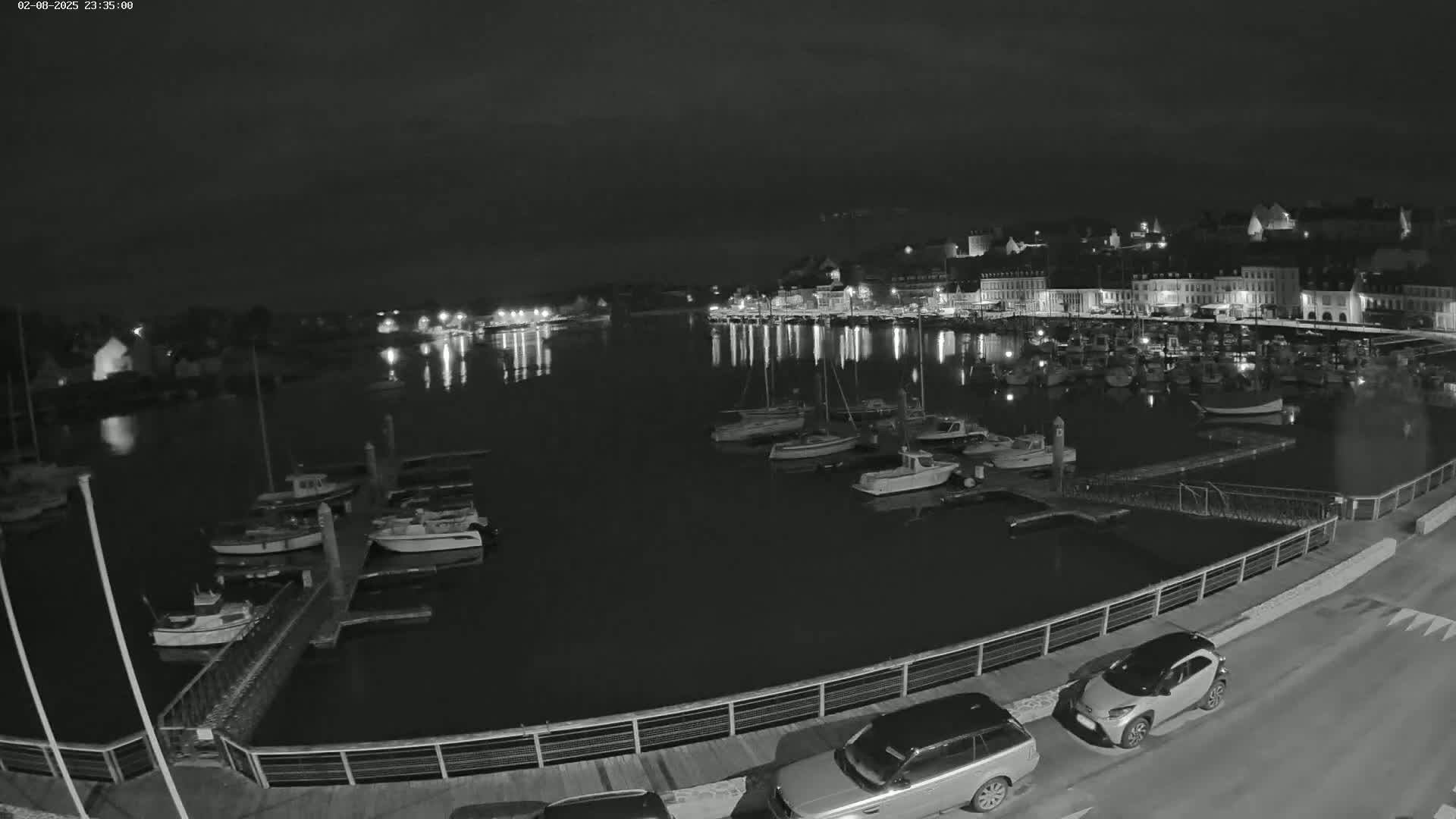 A nighttime, harbor view shows numerous boats docked along a waterfront, with a town's lights reflecting in the calm water.