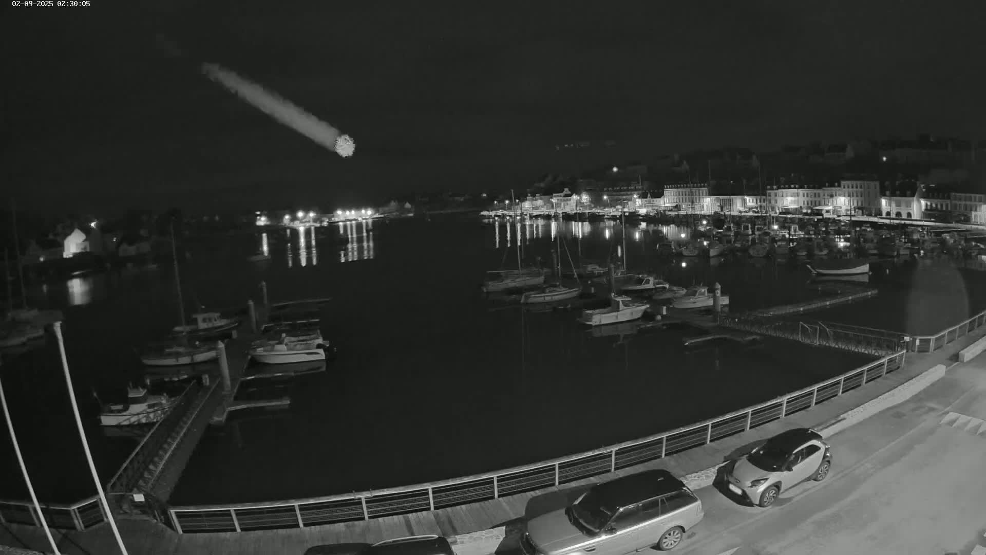 A nighttime, black and white view of a harbor filled with boats, a waterfront town, and a bright meteor streaking across the dark sky.