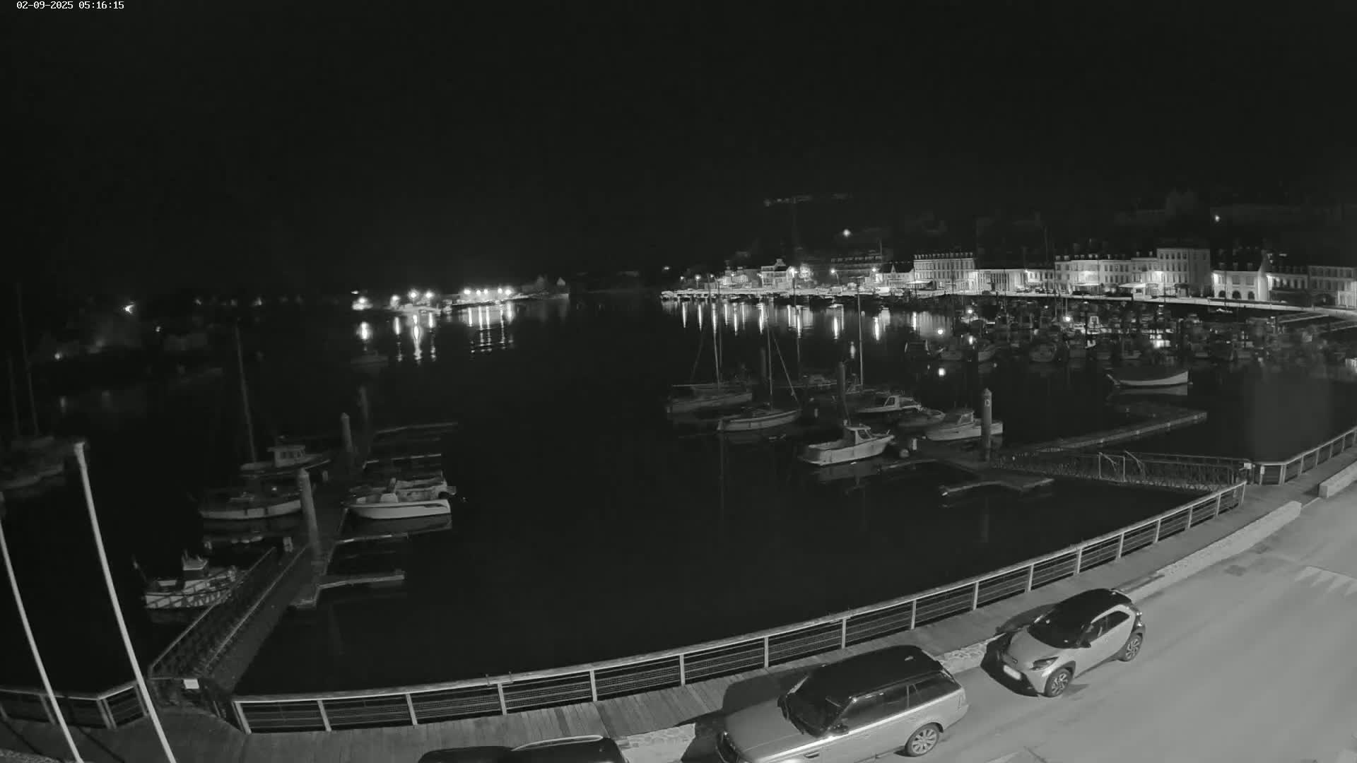 A nighttime view of a harbor filled with boats, docked alongside a waterfront town under a clear, dark sky.