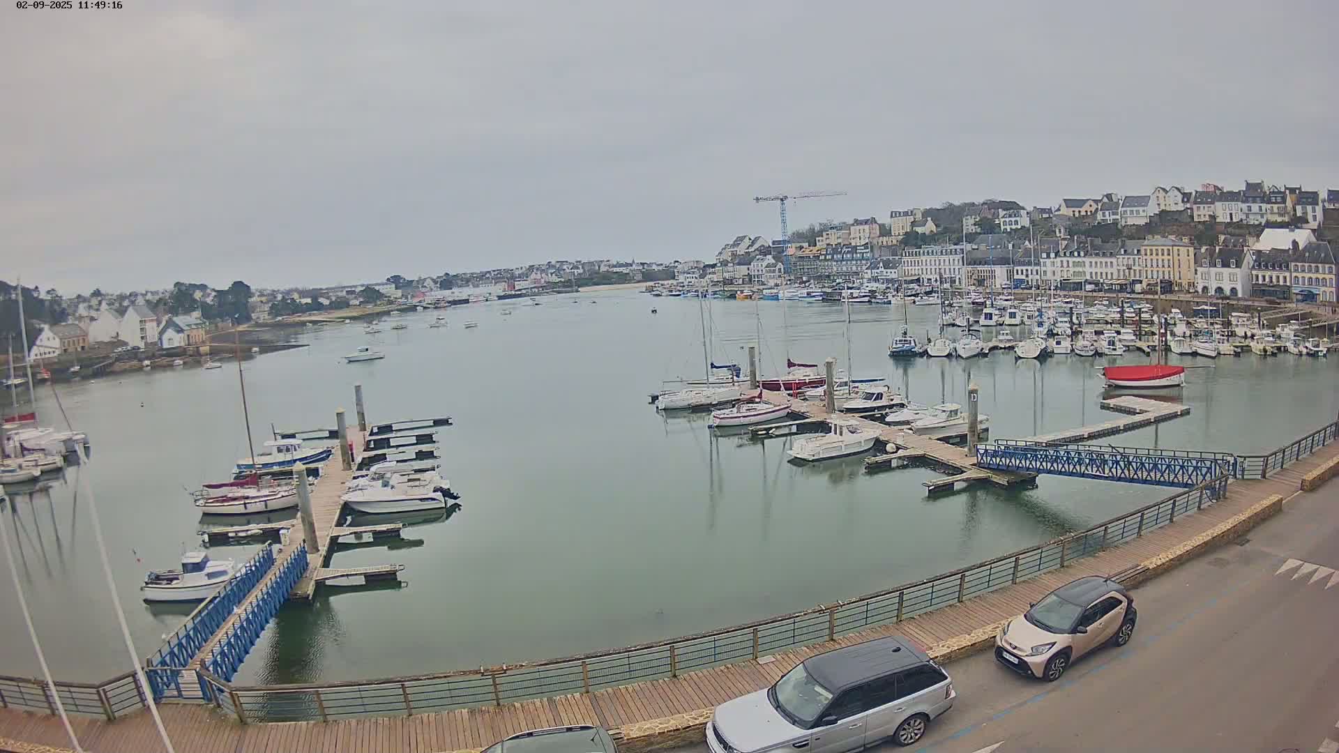 A harbor filled with numerous boats under an overcast sky, with a town and a construction crane visible in the background.