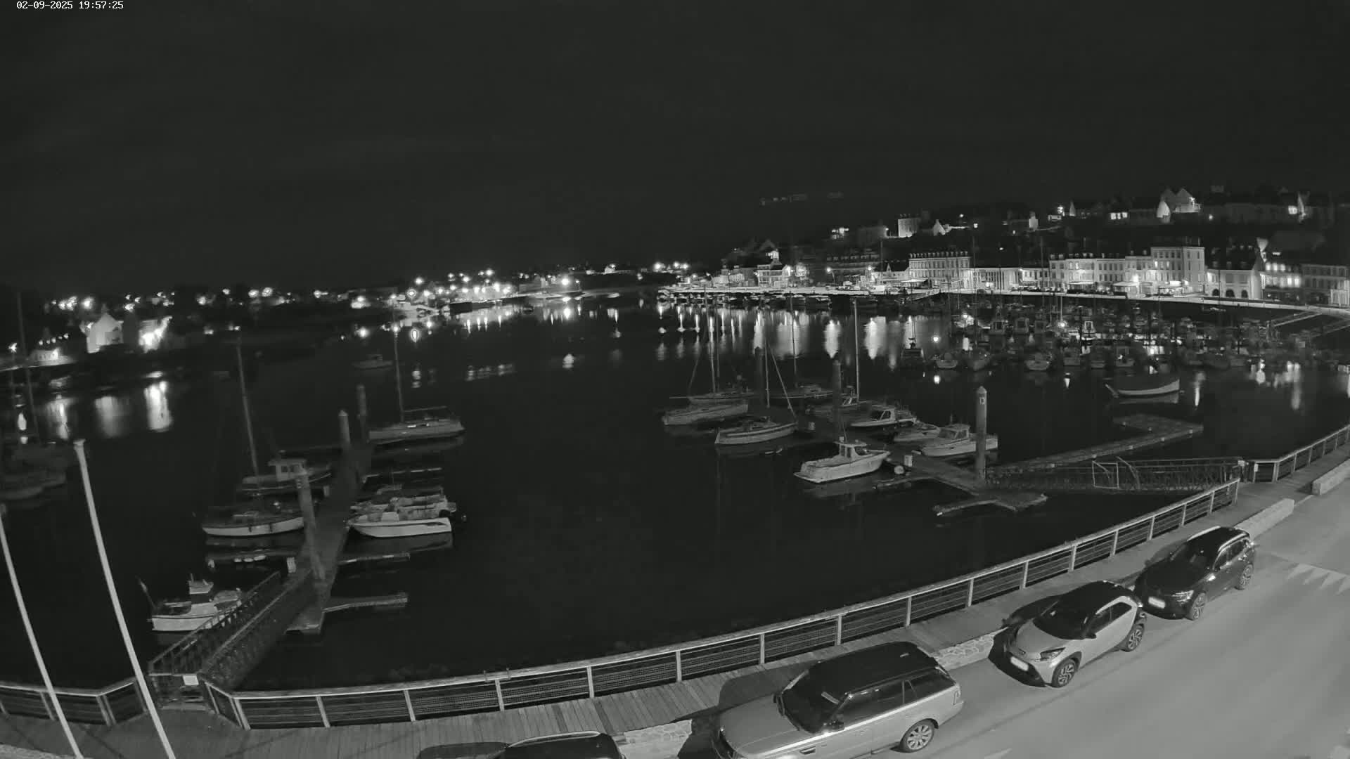 A nighttime, high-angle view of a harbor filled with boats, alongside a town with illuminated buildings, and several cars parked along a waterfront walkway.