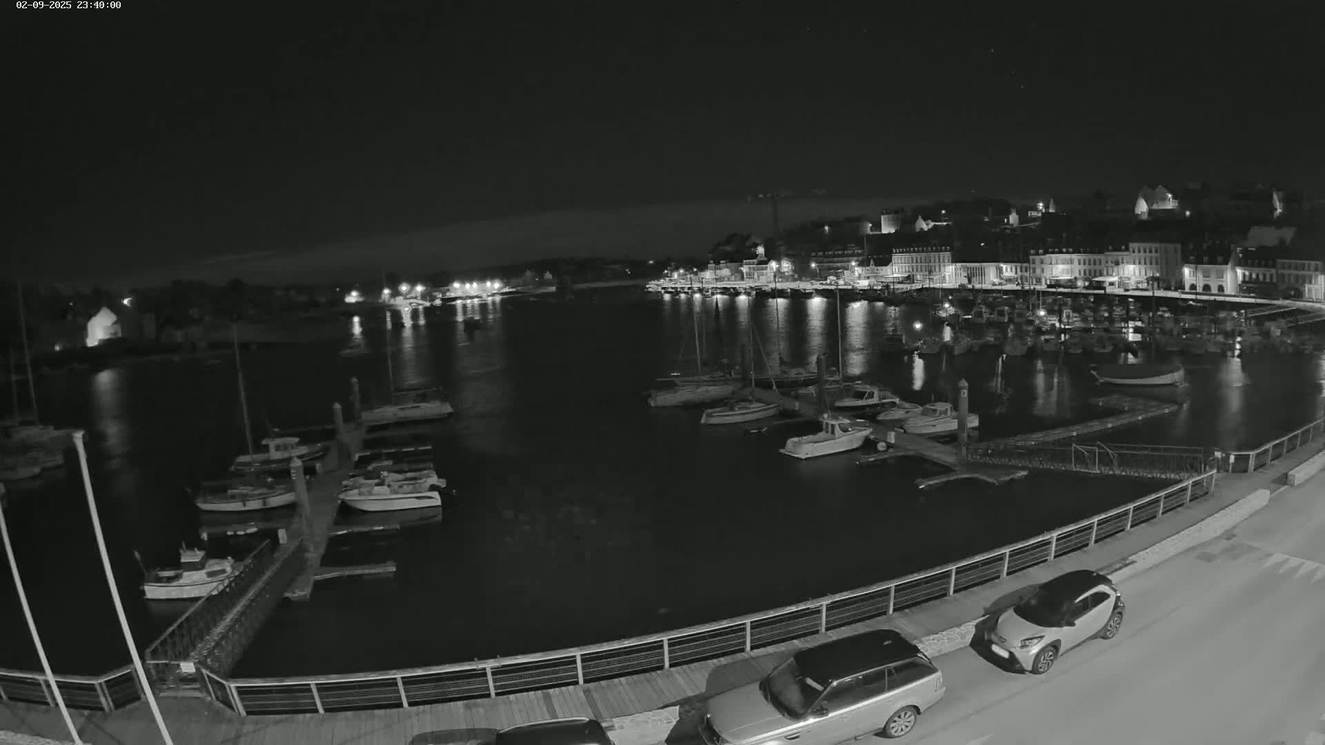 A nighttime view of a harbor filled with boats, a town with lights reflecting on the water, and several cars parked along a waterfront walkway under a clear, dark sky.