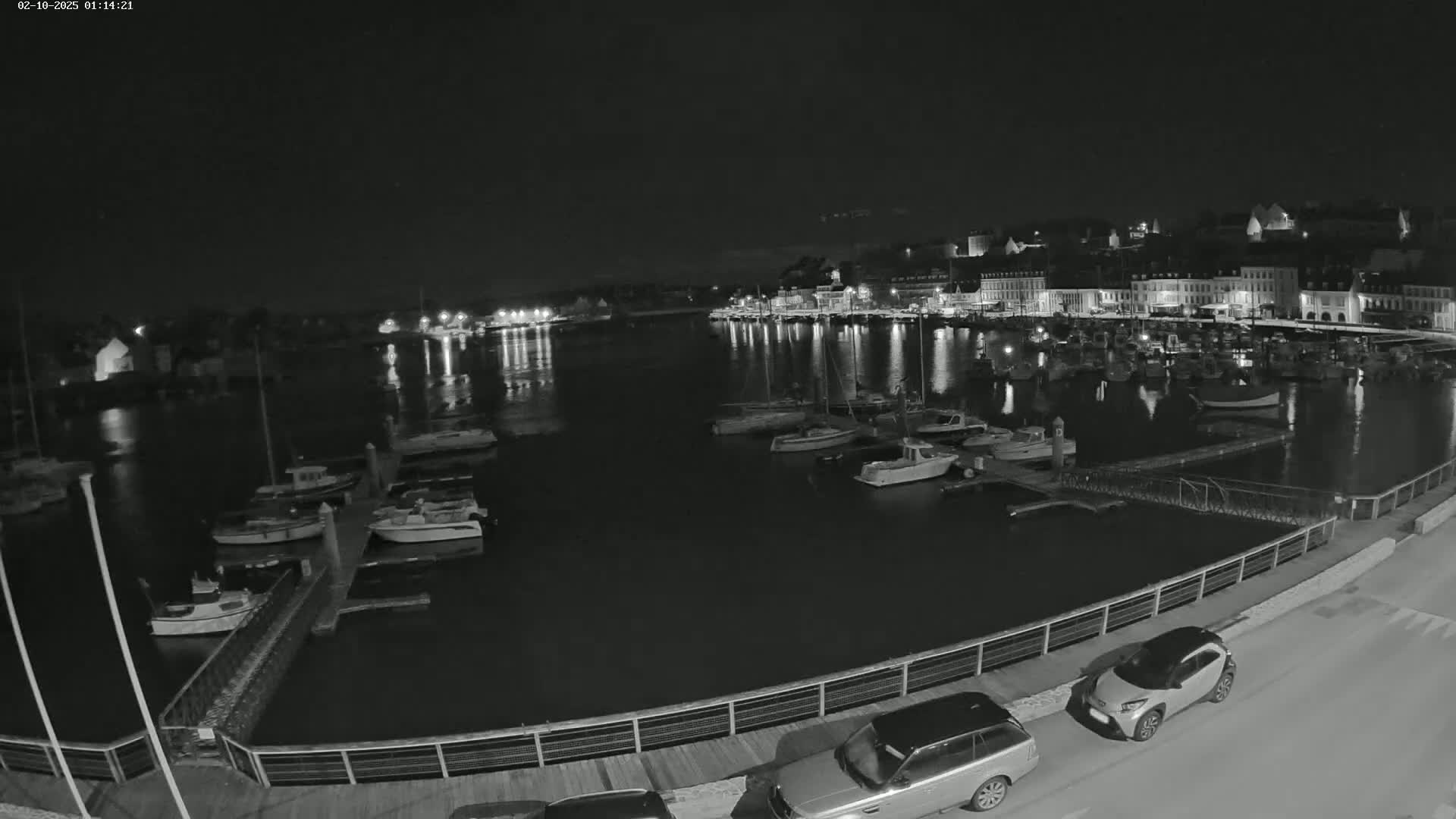 A nighttime, black-and-white view of a harbor filled with boats, a waterfront town with illuminated buildings, and several cars parked along a walkway.