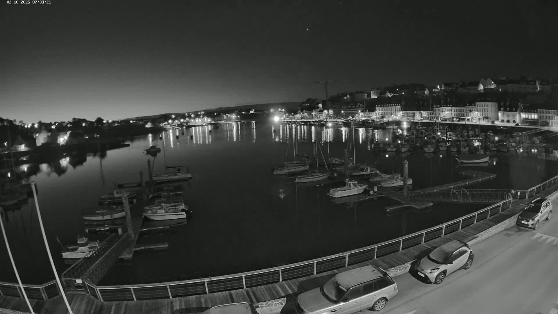 A nighttime, black-and-white view of a harbor filled with boats, alongside a waterfront with parked cars and buildings lit up in the background.