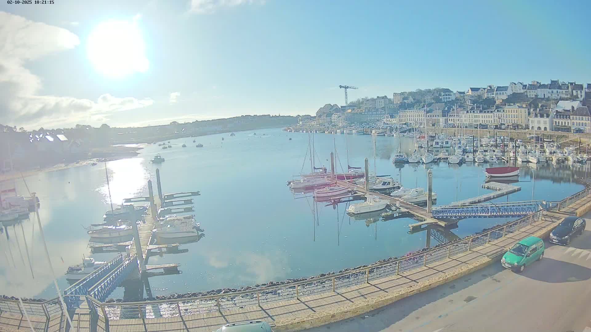 A sunny day shows a harbor filled with numerous boats docked at piers, with a town and a construction crane visible in the background.