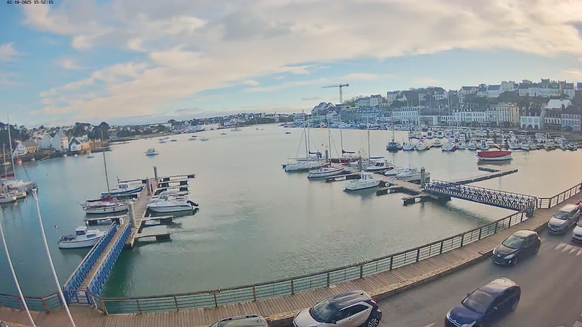 A harbor filled with sailboats and motorboats under a partly cloudy sky is viewed from a boardwalk with several cars parked nearby.