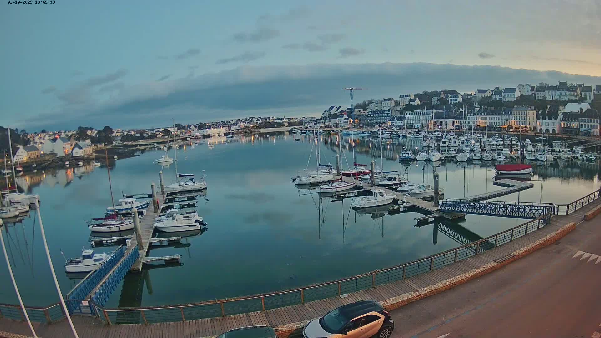 A calm harbor filled with numerous boats at dusk, under a cloudy sky, is viewed from a slightly elevated position.