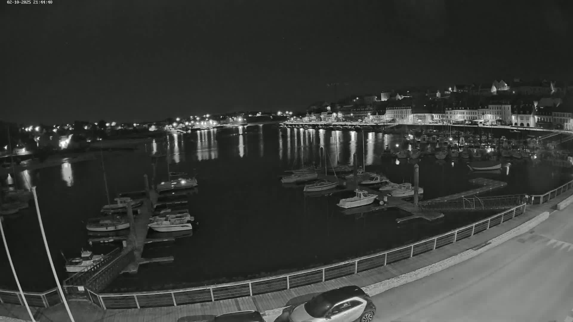 A nighttime view of a calm harbor filled with docked boats, reflecting the lights of a town on the opposite shore.