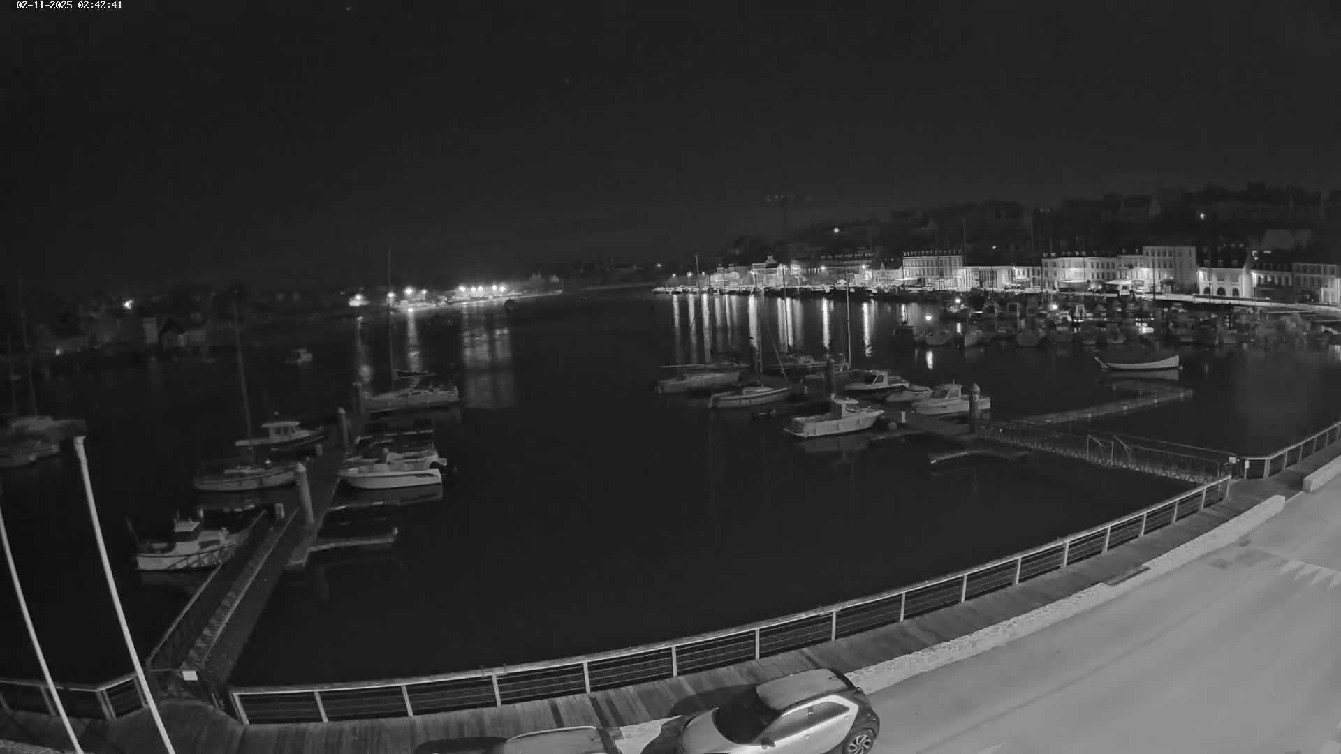 A nighttime view of a harbor filled with boats and docked along a waterfront, illuminated by building lights under a clear, dark sky.