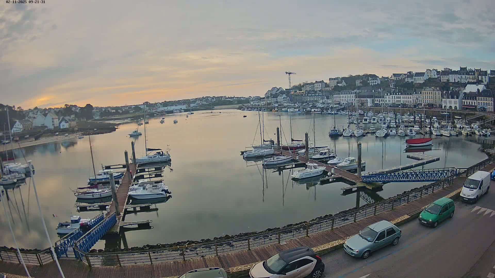 A calm harbor filled with numerous sailboats and motorboats at sunrise, under a mostly clear sky, is overlooked by a town of white buildings.