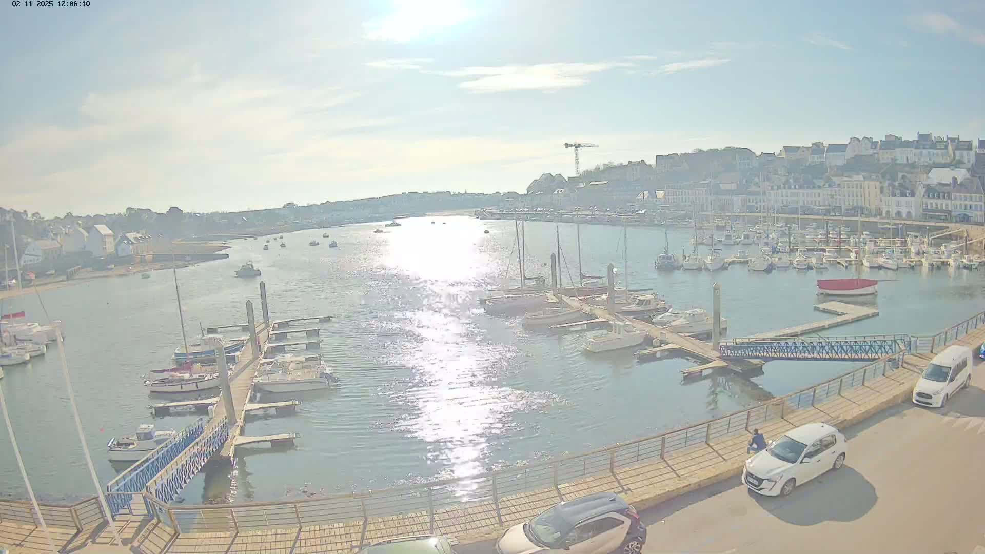 A sunny harbor scene shows numerous boats docked at piers, with a town and buildings visible in the background.