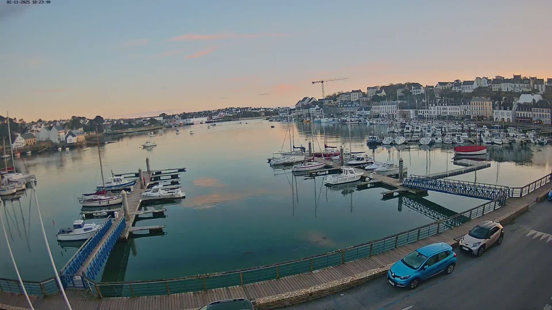 A calm harbor filled with numerous sailboats and motorboats at sunset, under a clear, pale pink sky.