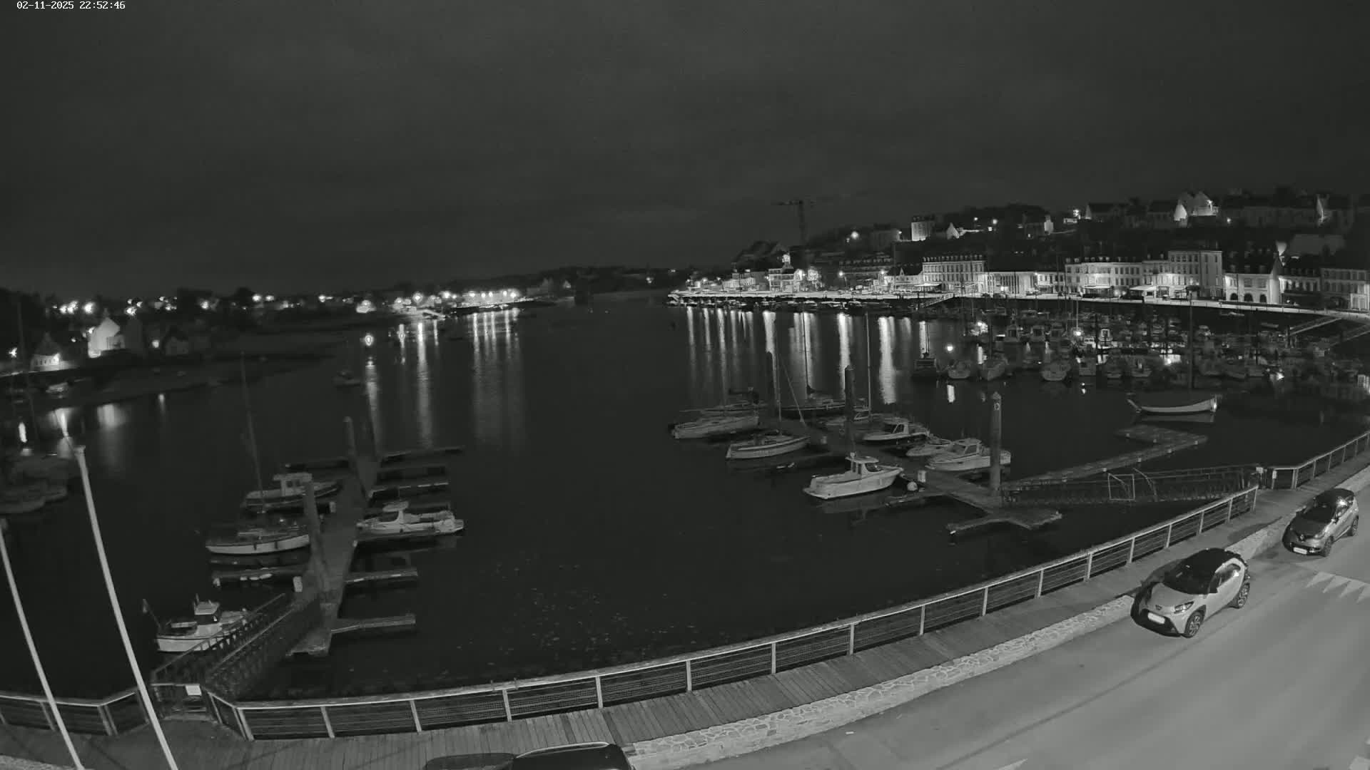 A nighttime, grayscale view of a harbor filled with docked boats, with a town's lights reflected in the calm water.