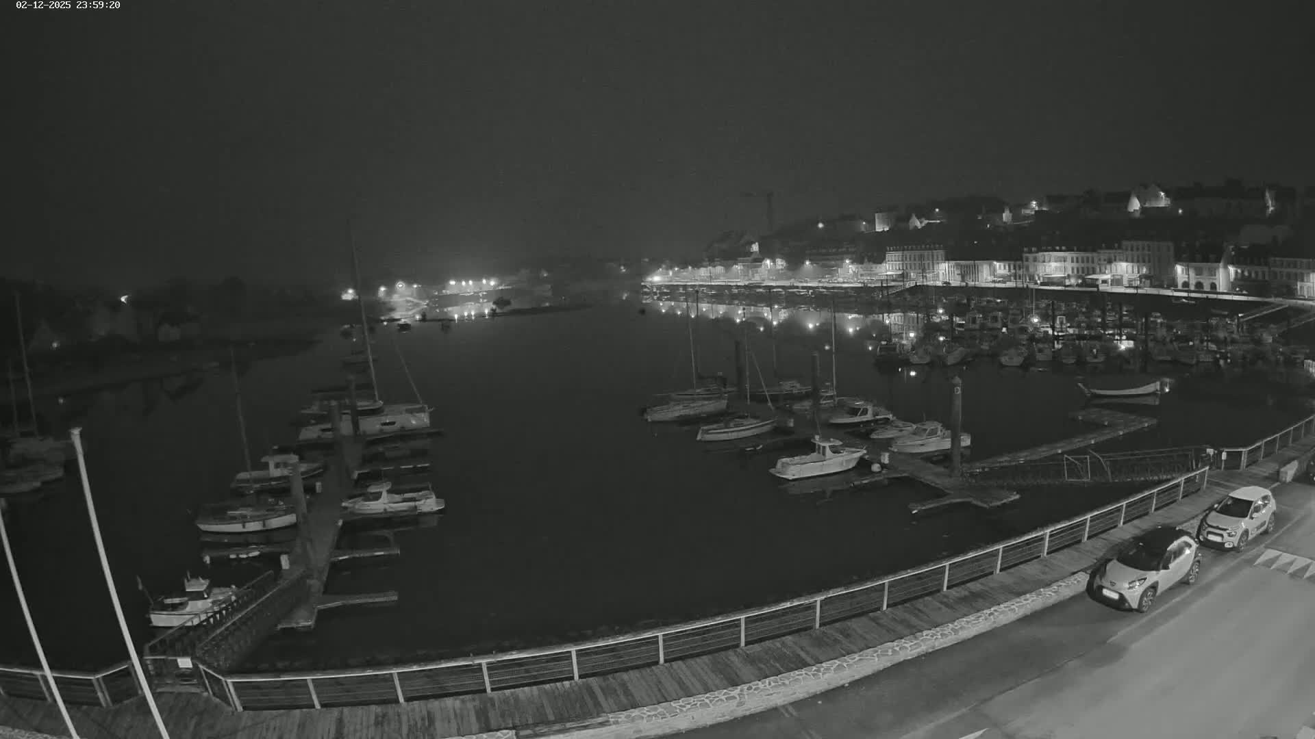 A nighttime view of a harbor filled with docked boats, a town's waterfront buildings illuminated, and two cars parked along a nearby walkway under a dark, possibly foggy sky.