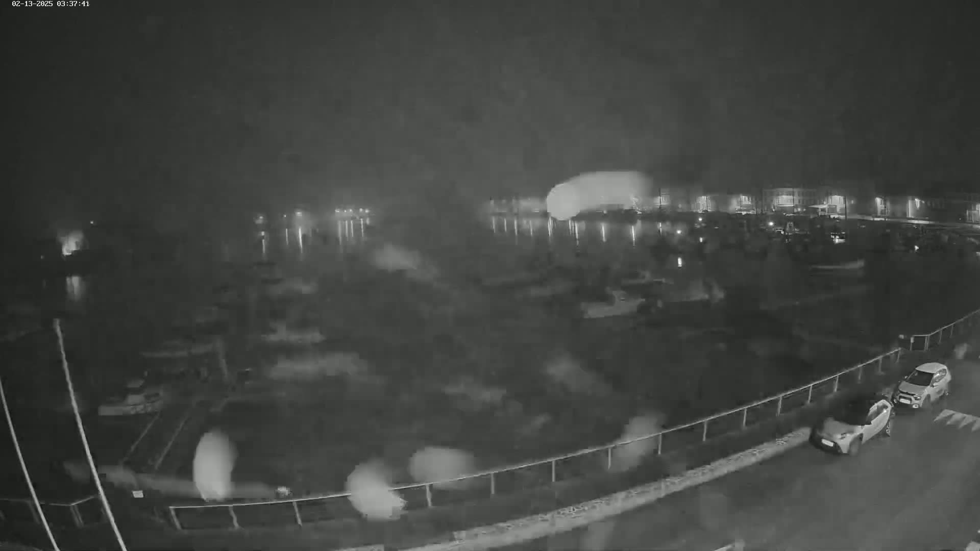 A nighttime, foggy view of a harbor with several parked cars along a roadway and buildings faintly visible in the background.
