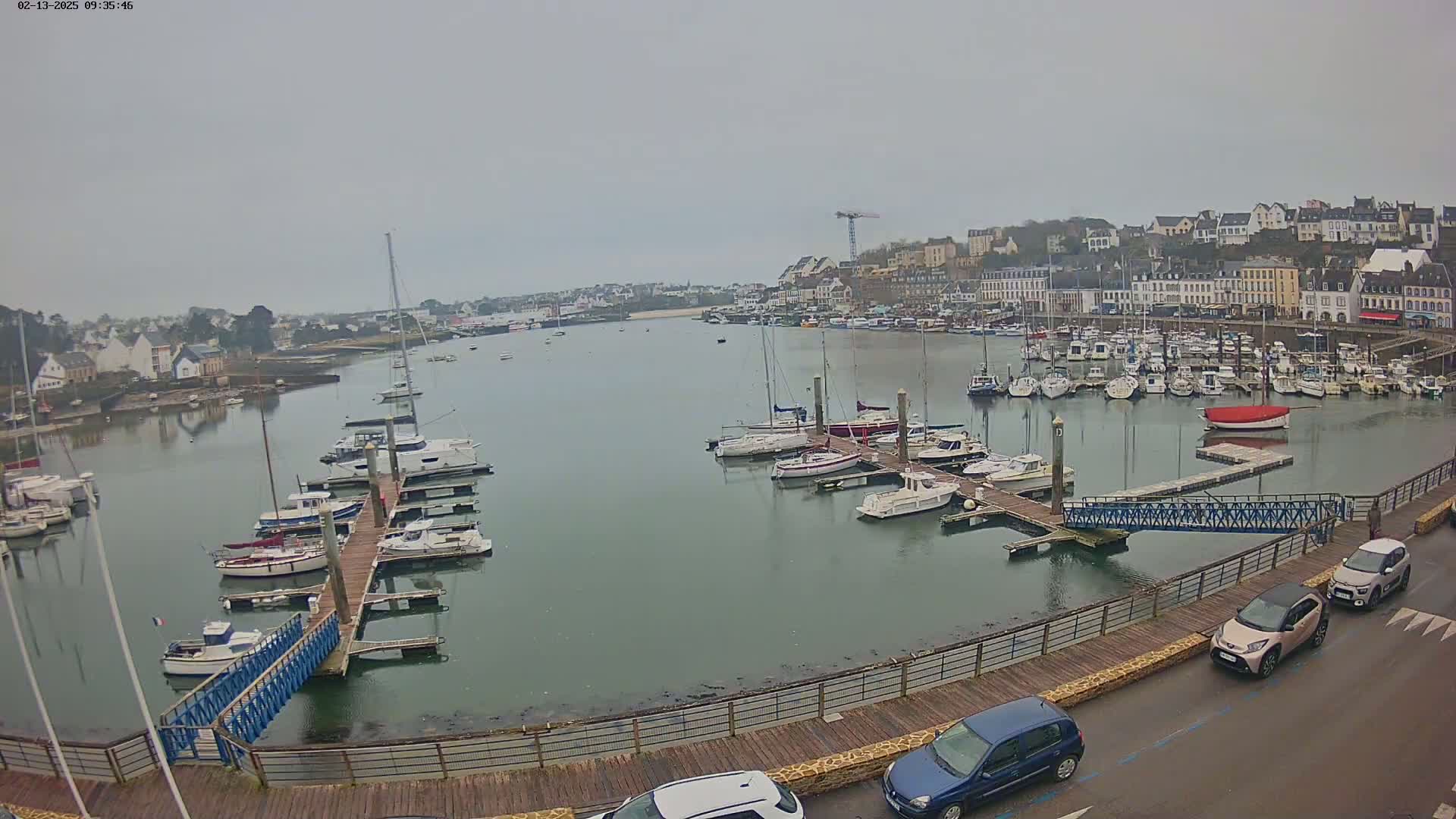 A harbor filled with numerous sailboats and motorboats docked at piers on a cloudy day, with a town visible in the background.