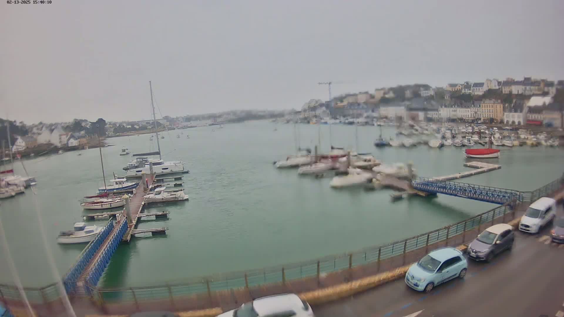 A harbor filled with numerous boats and yachts on a cloudy day, viewed from a slightly elevated perspective overlooking a roadway with several cars parked.