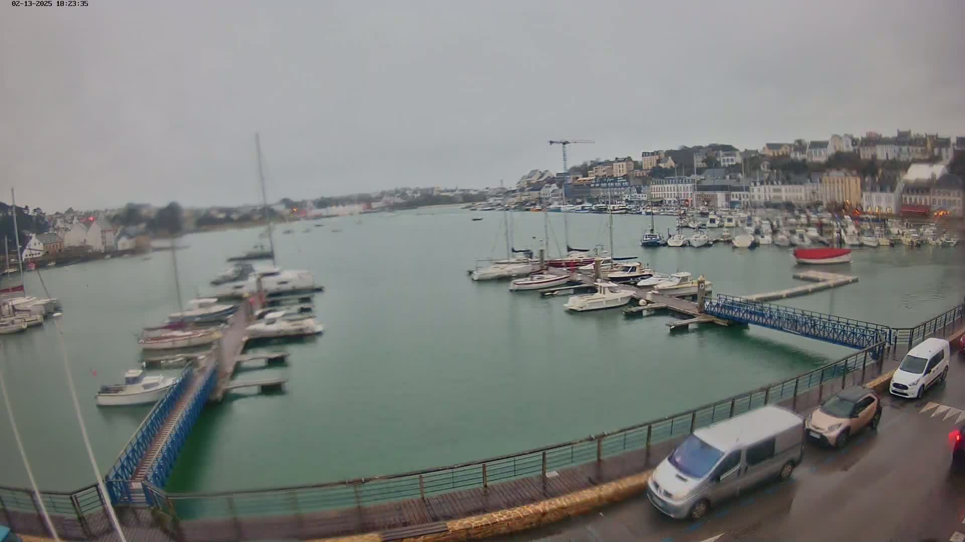 A harbor filled with numerous boats on a cloudy day, viewed from a waterfront promenade with several vehicles parked along the side.