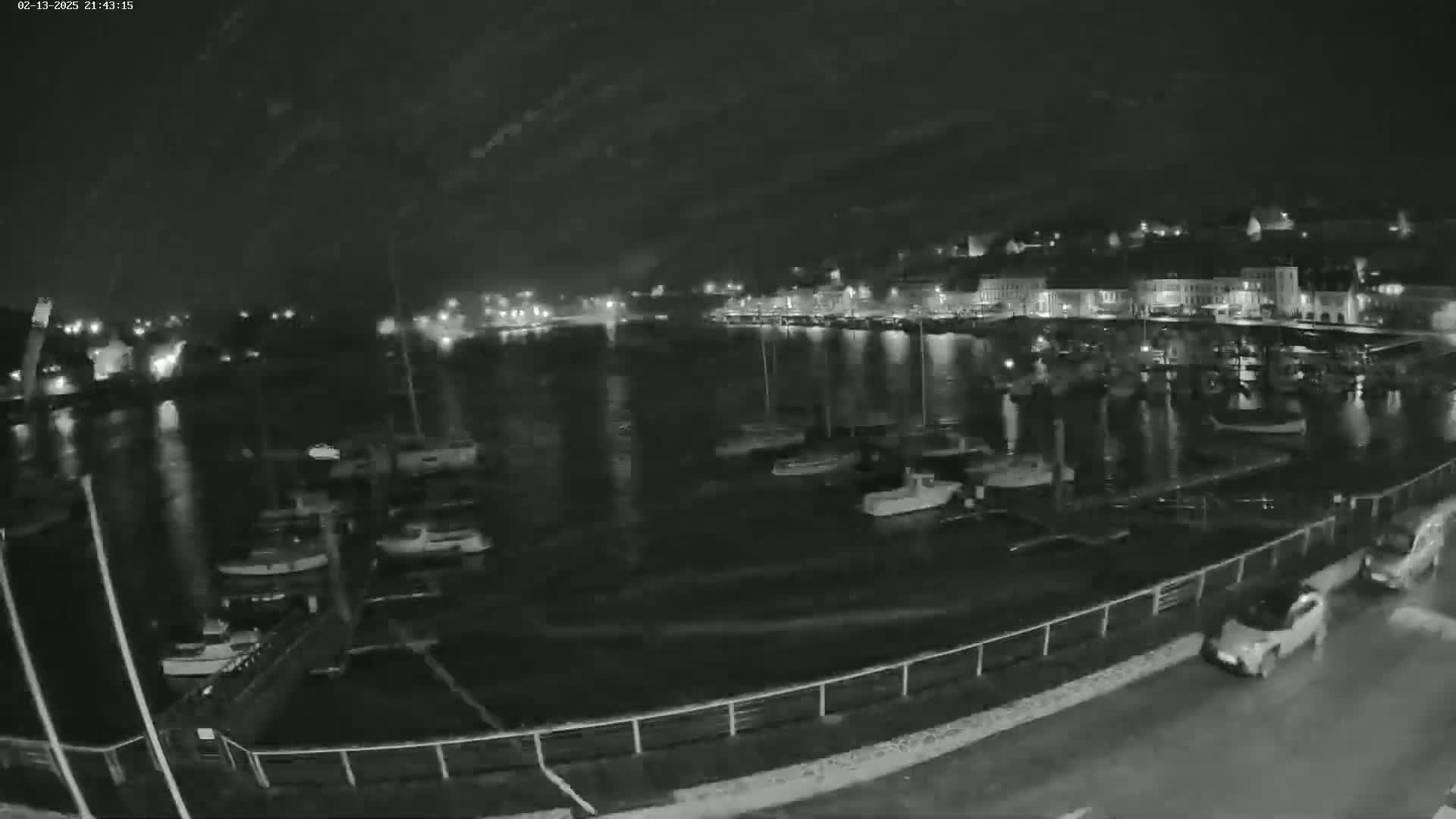 A nighttime, low-light view of a harbor filled with boats, with a town's lights visible across the water.