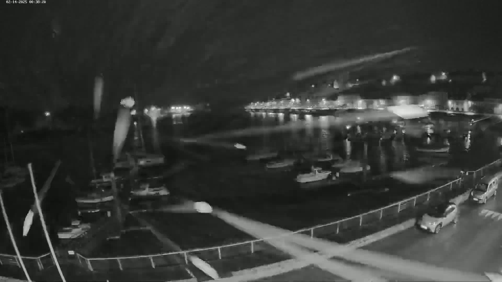 A nighttime, low-light view of a harbor with several boats and cars parked along a waterfront, with rain or snow falling.