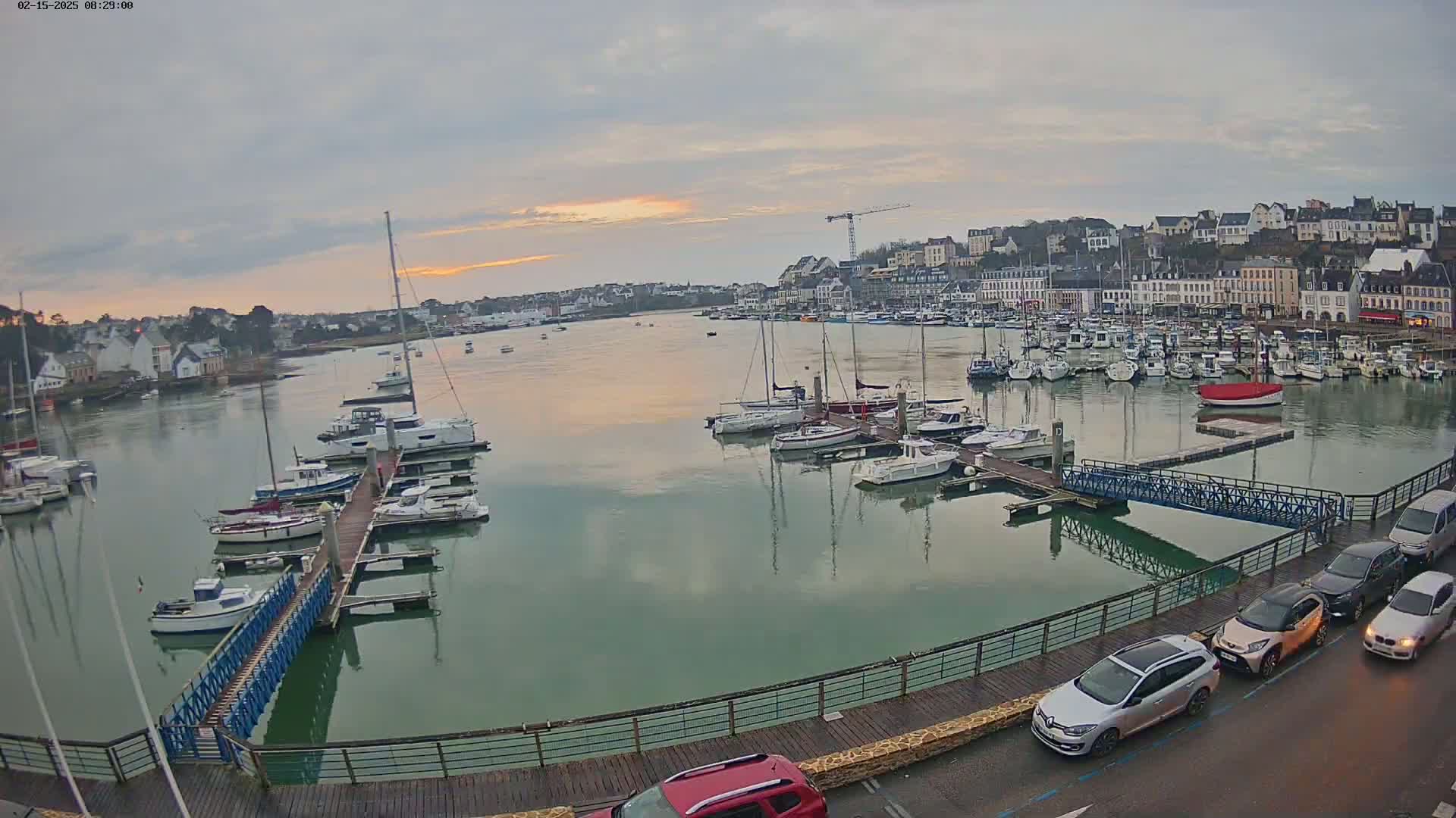 A calm harbor filled with sailboats and motorboats at dawn under an overcast sky, with a town and cars visible along the shoreline.