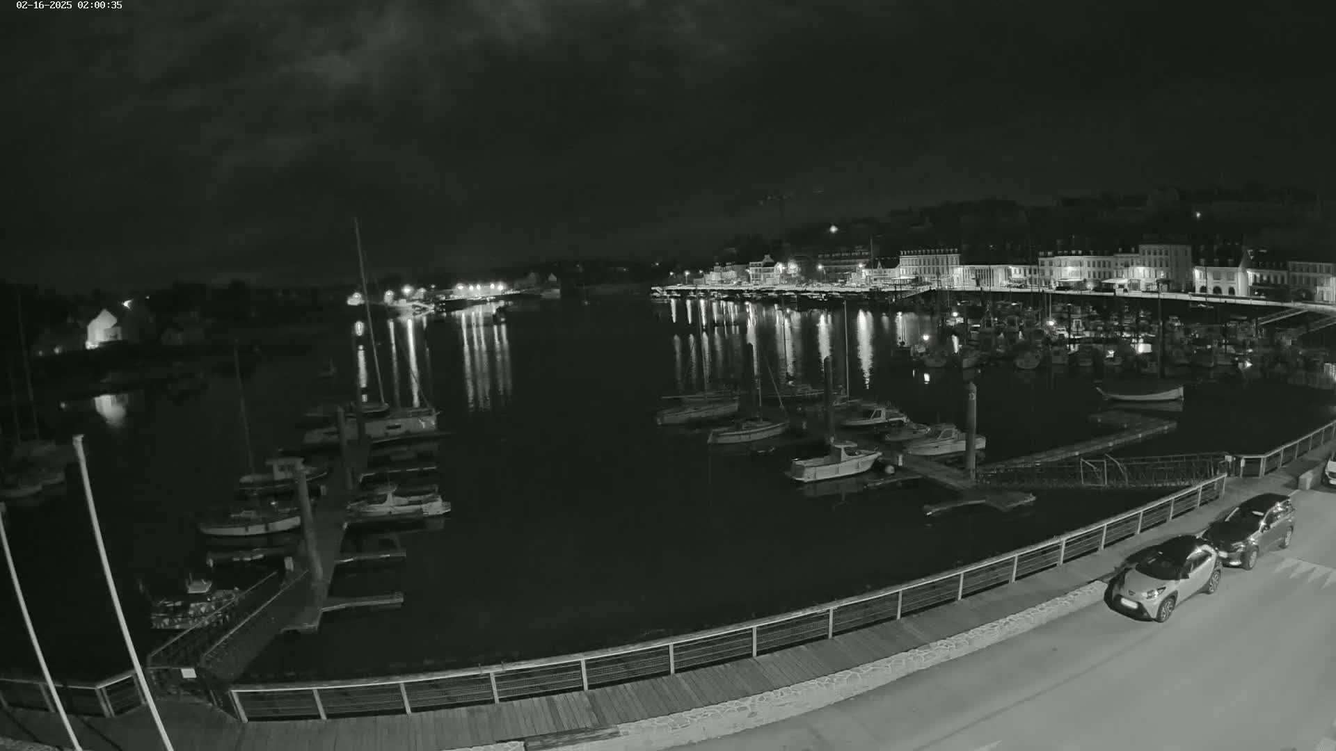 A nighttime view of a harbor filled with boats, docked along a waterfront with buildings and lights across the water under a mostly cloudy sky.