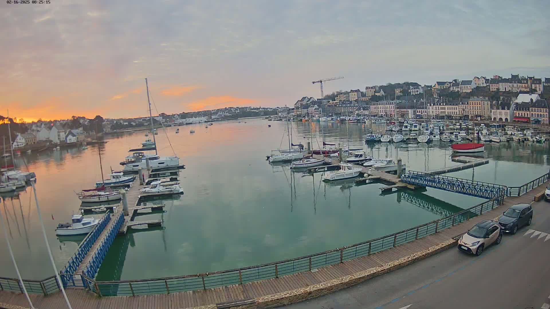 A calm harbor filled with sailboats and motorboats at sunrise, overlooked by a town with multi-story buildings.