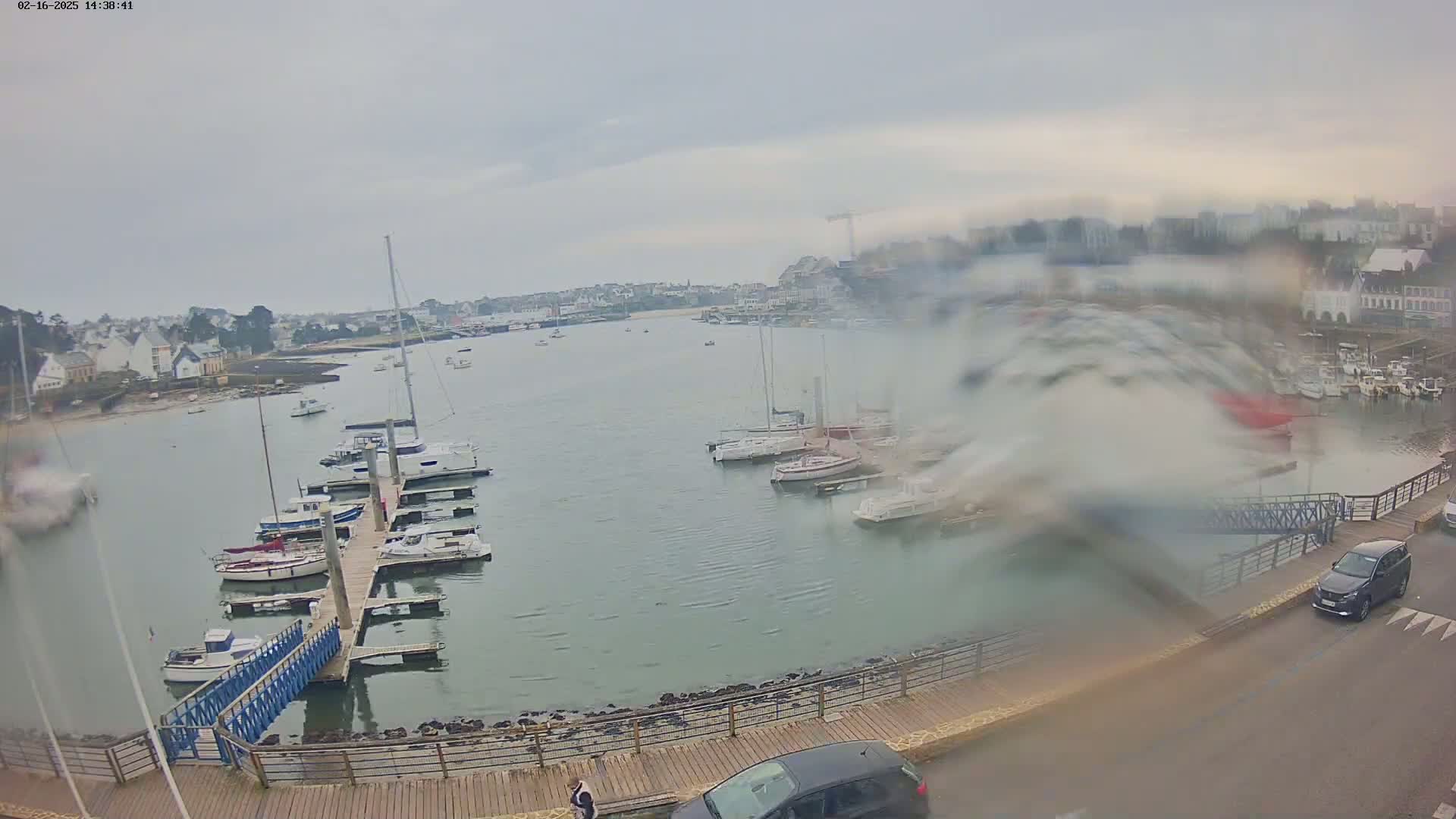 A harbor filled with boats on a rainy day, viewed from an elevated position overlooking a waterfront walkway and parked cars.