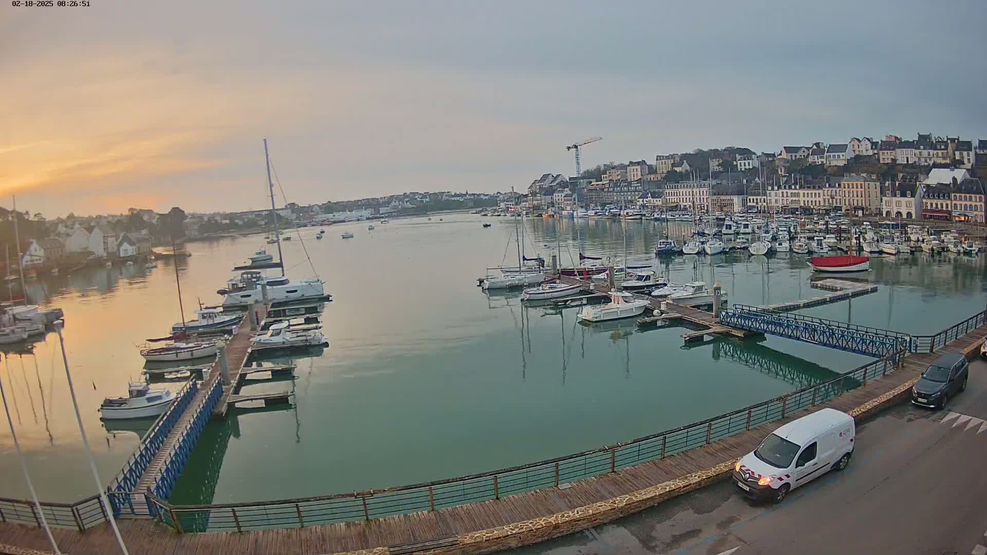 A calm harbor filled with numerous sailboats and motorboats at sunrise, overlooked by a town with buildings lining the waterfront.