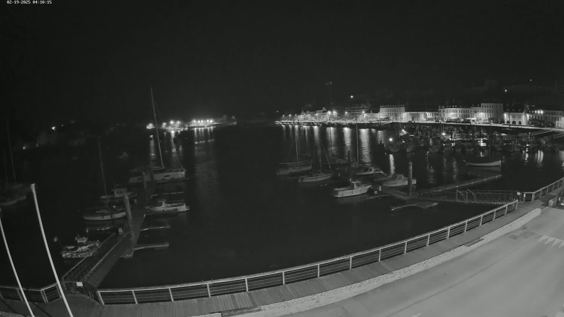 A nighttime, black-and-white view of a harbor filled with numerous boats moored alongside a waterfront lined with buildings.