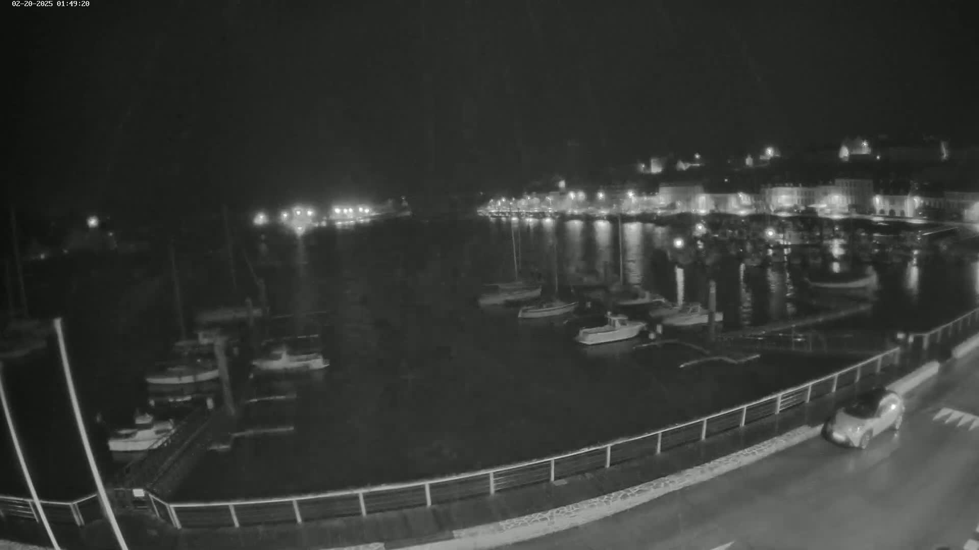 A nighttime view of a harbor filled with boats, a town with lights visible across the water, and a car driving along a wet road next to a railing.