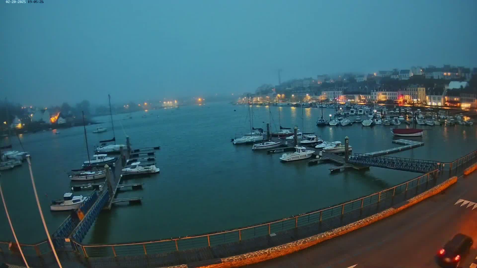 A harbor filled with docked sailboats is shrouded in a misty twilight, with a town's lights visible in the distance.
