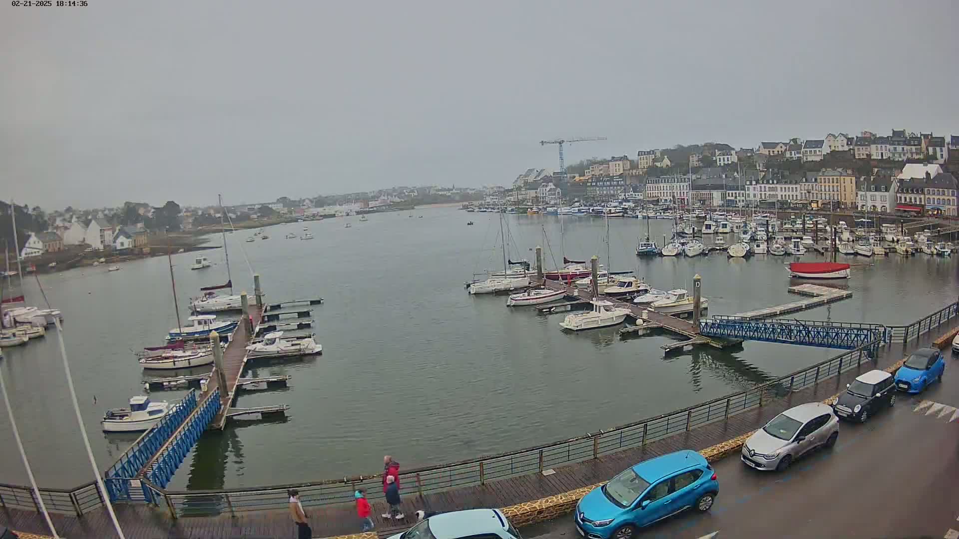 A harbor filled with numerous boats on a cloudy day, with a town in the background and several people walking along a boardwalk.